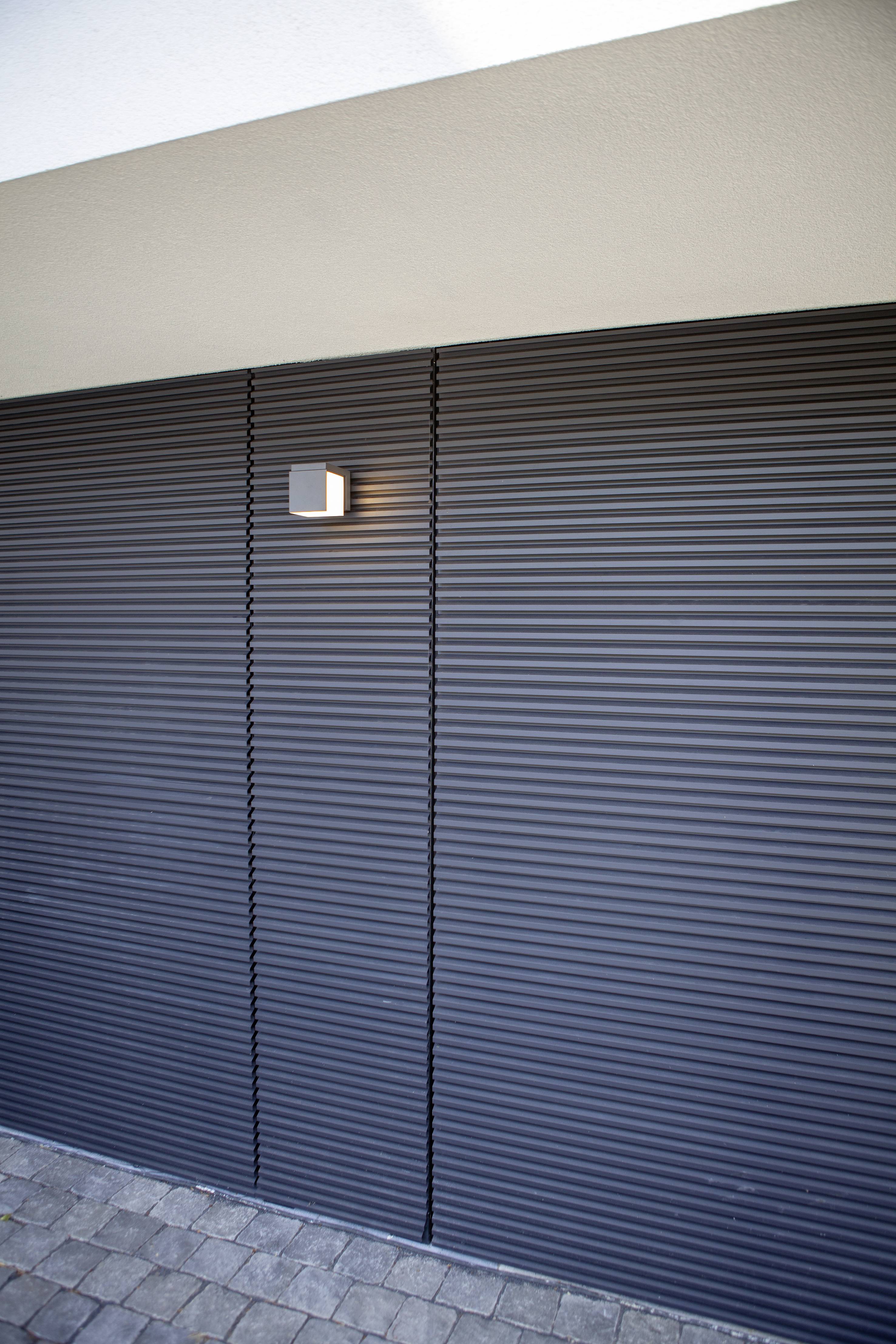 Porte de garage gris anthracite avec rainures verticales, éclairée par une petite applique murale située au-dessus à gauche, sur un sol pavé.