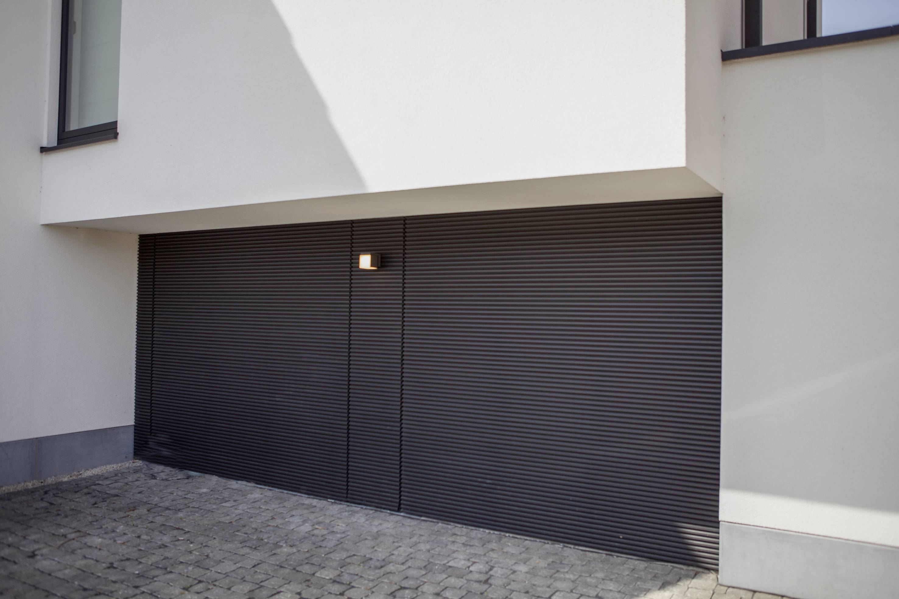 Une porte de garage fermée sous un bâtiment moderne et blanc. L'entrée paraît minimaliste et épurée.