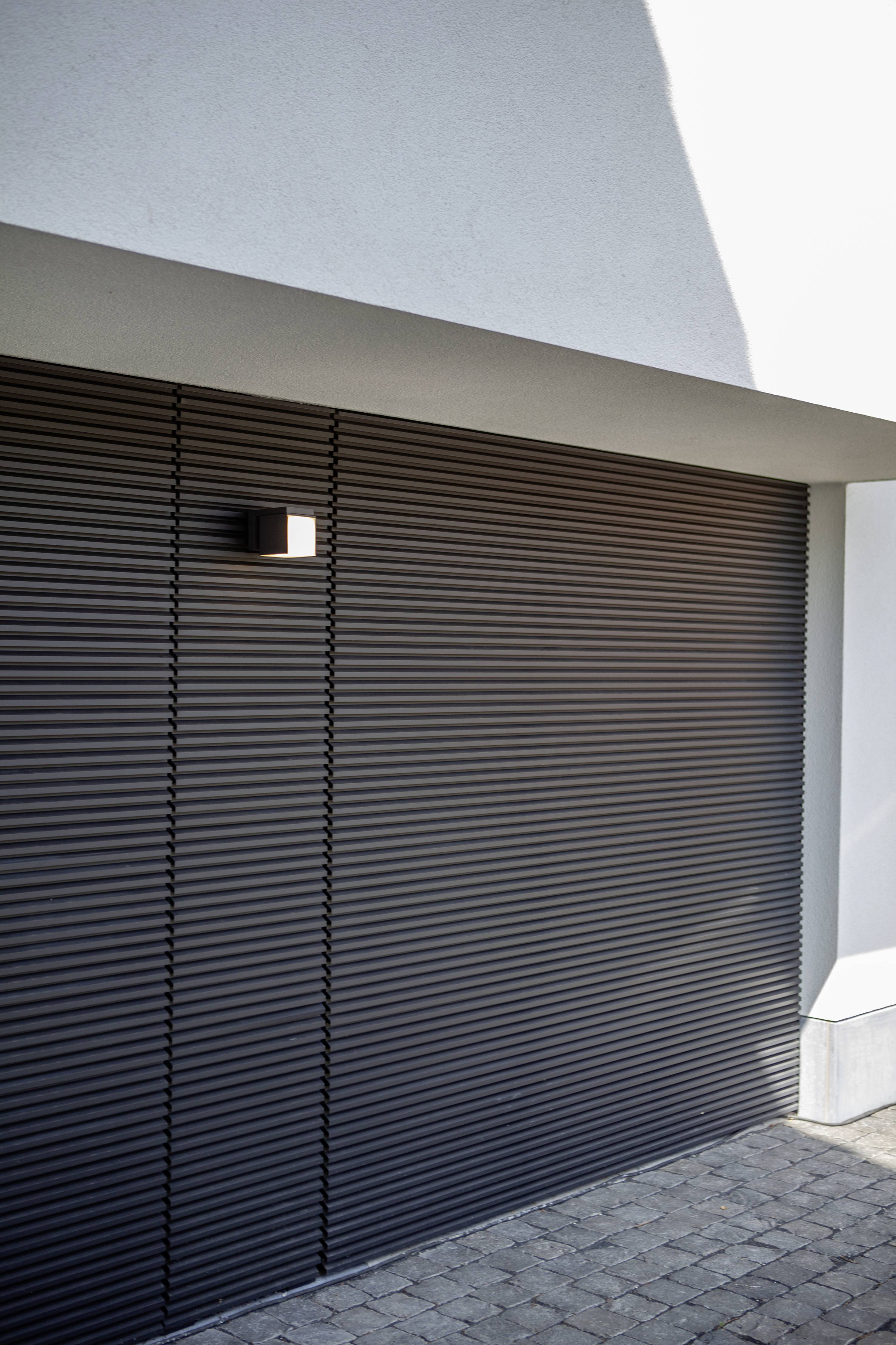 Porte de garage grise fermée avec une structure horizontale rainurée moderne. Ombre projetée sur le mur, pavés en premier plan.