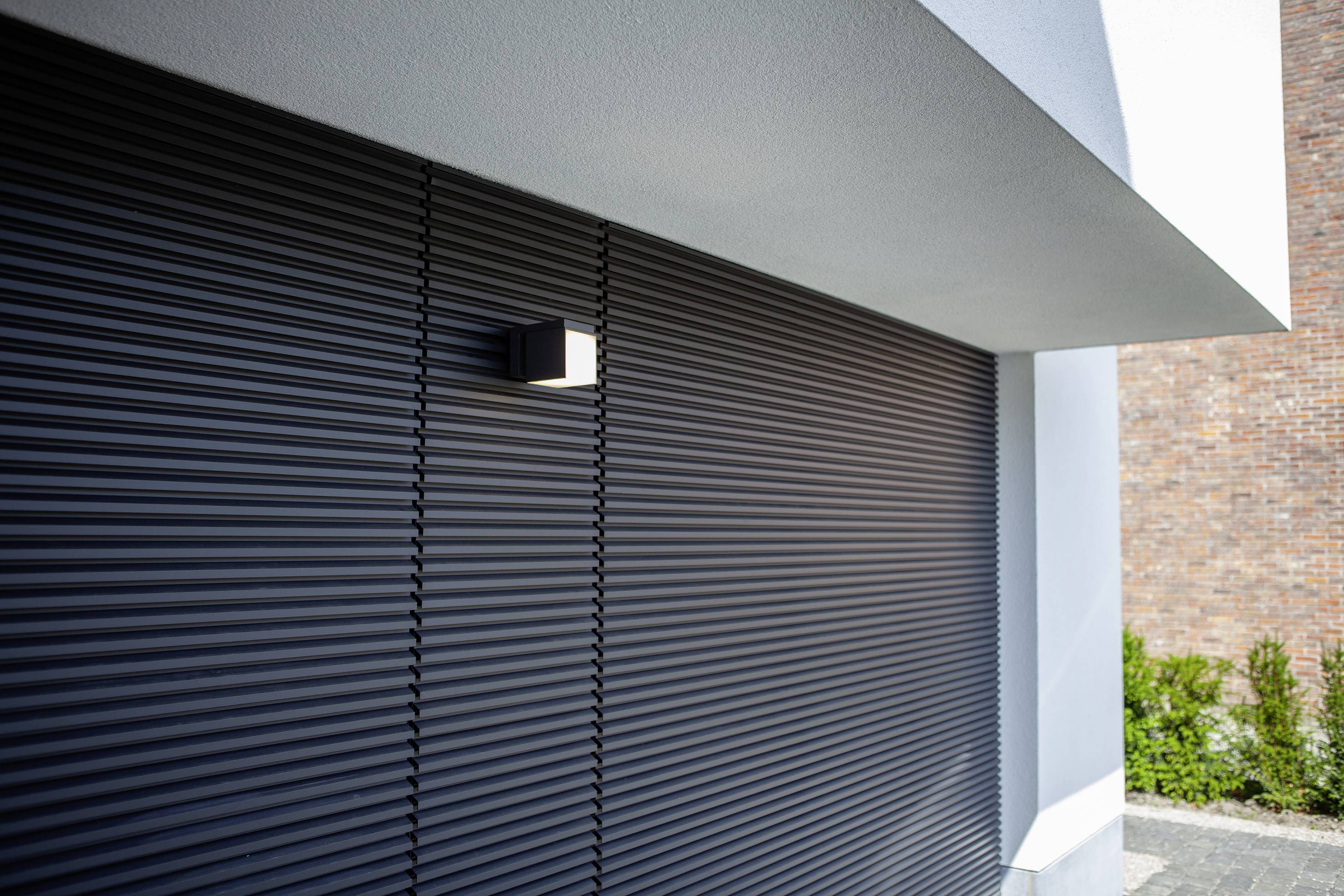 Porte de garage avec des lamelles sombres, architecture moderne avec un auvent blanc, petit cube de lampe murale, mur de briques et plantes en arrière-plan.