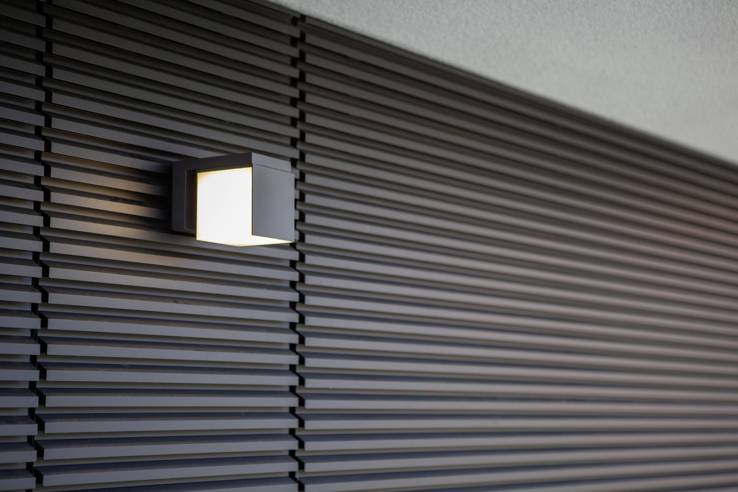 Une lampe extérieure moderne et rectangulaire éclaire un mur gris ondulé.
