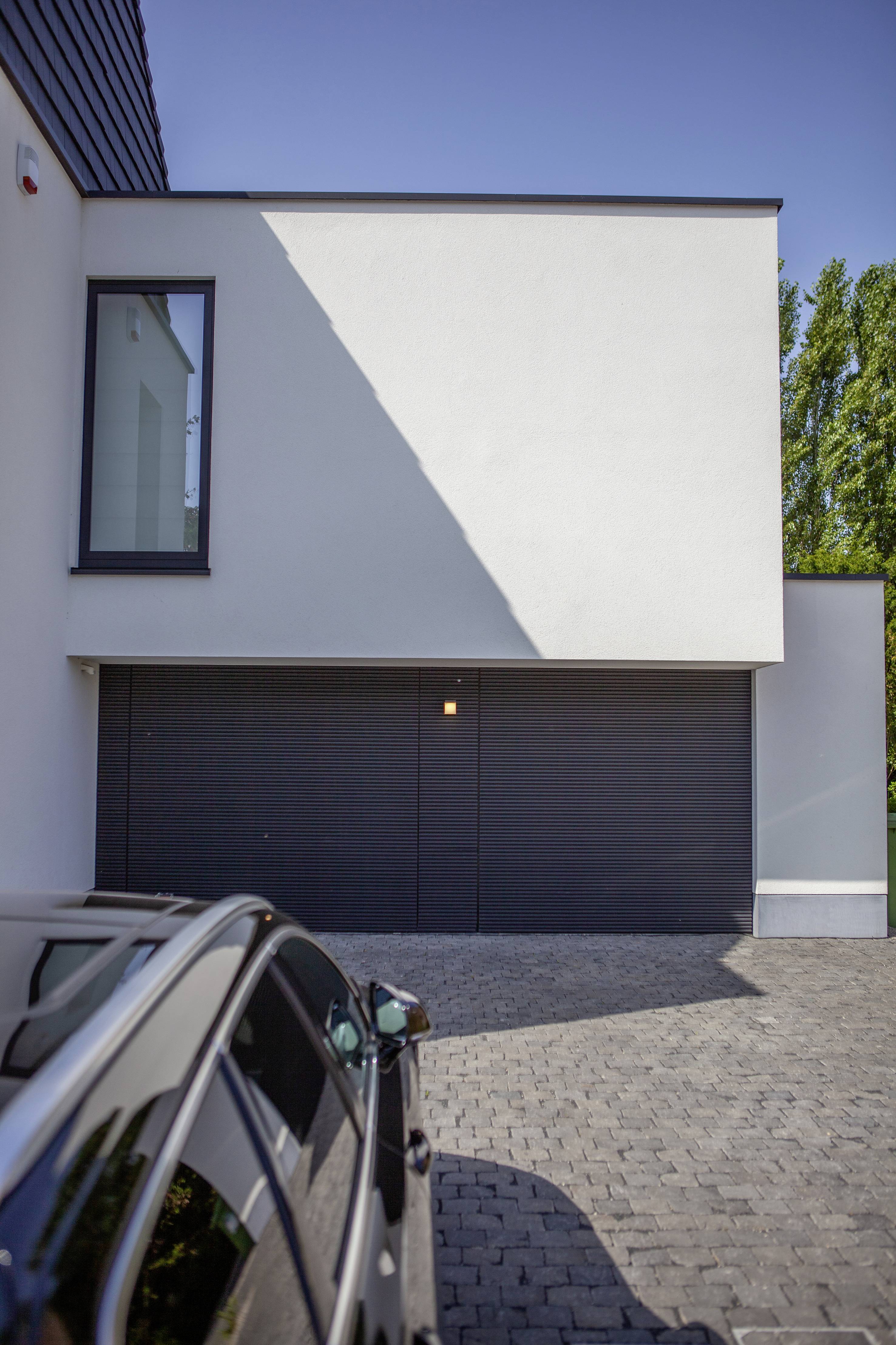 Une maison individuelle moderne, blanche, avec une grande fenêtre et des portes de garage sombres. Au premier plan, on aperçoit une voiture garée.