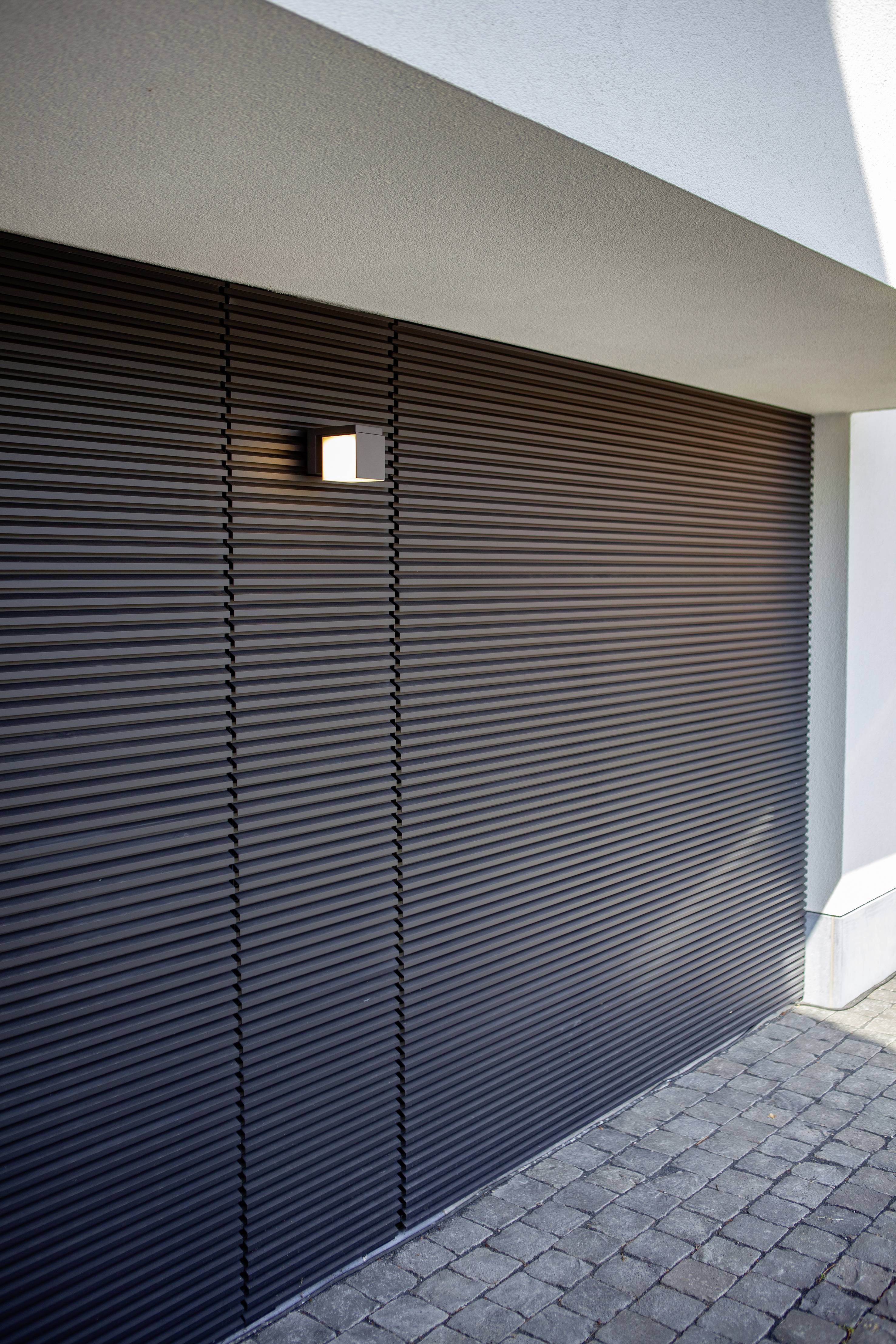 Un garage moderne avec une porte de garage noire à enroulement et une petite fenêtre carrée à côté d'un chemin pavé.