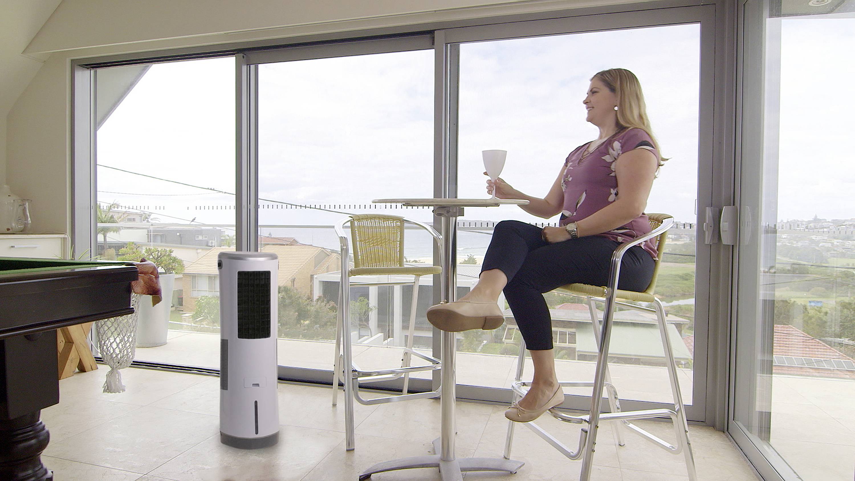 Une femme est assise à une table dans une pièce avec de grandes fenêtres et savoure une boisson. À côté d'elle se trouve un climatiseur portable.