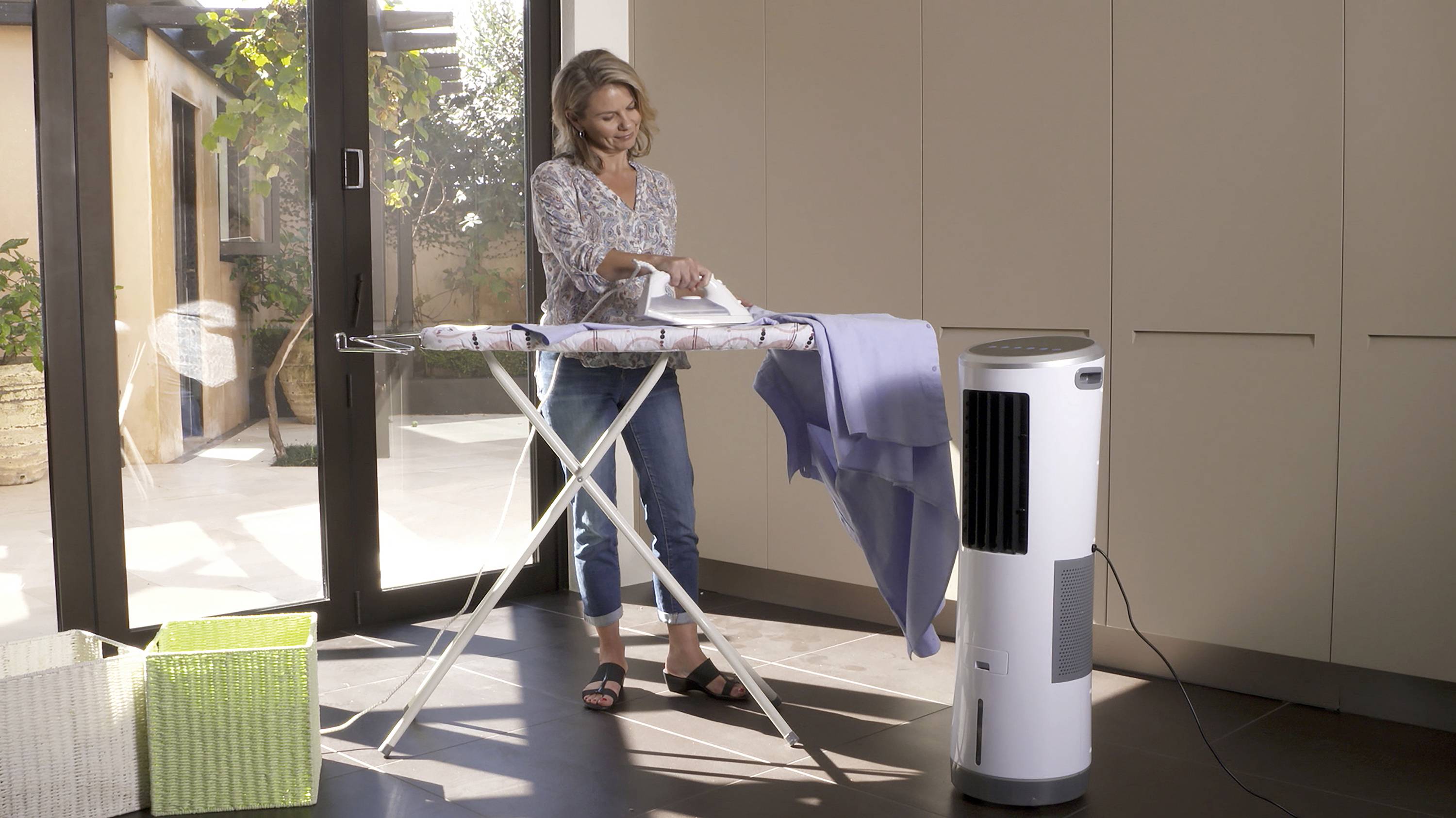 Une femme repasse une chemise bleue dans une pièce lumineuse, à côté d'un ventilateur. Elle porte un jean et un chemisier. Un grand placard se trouve en arrière-plan.