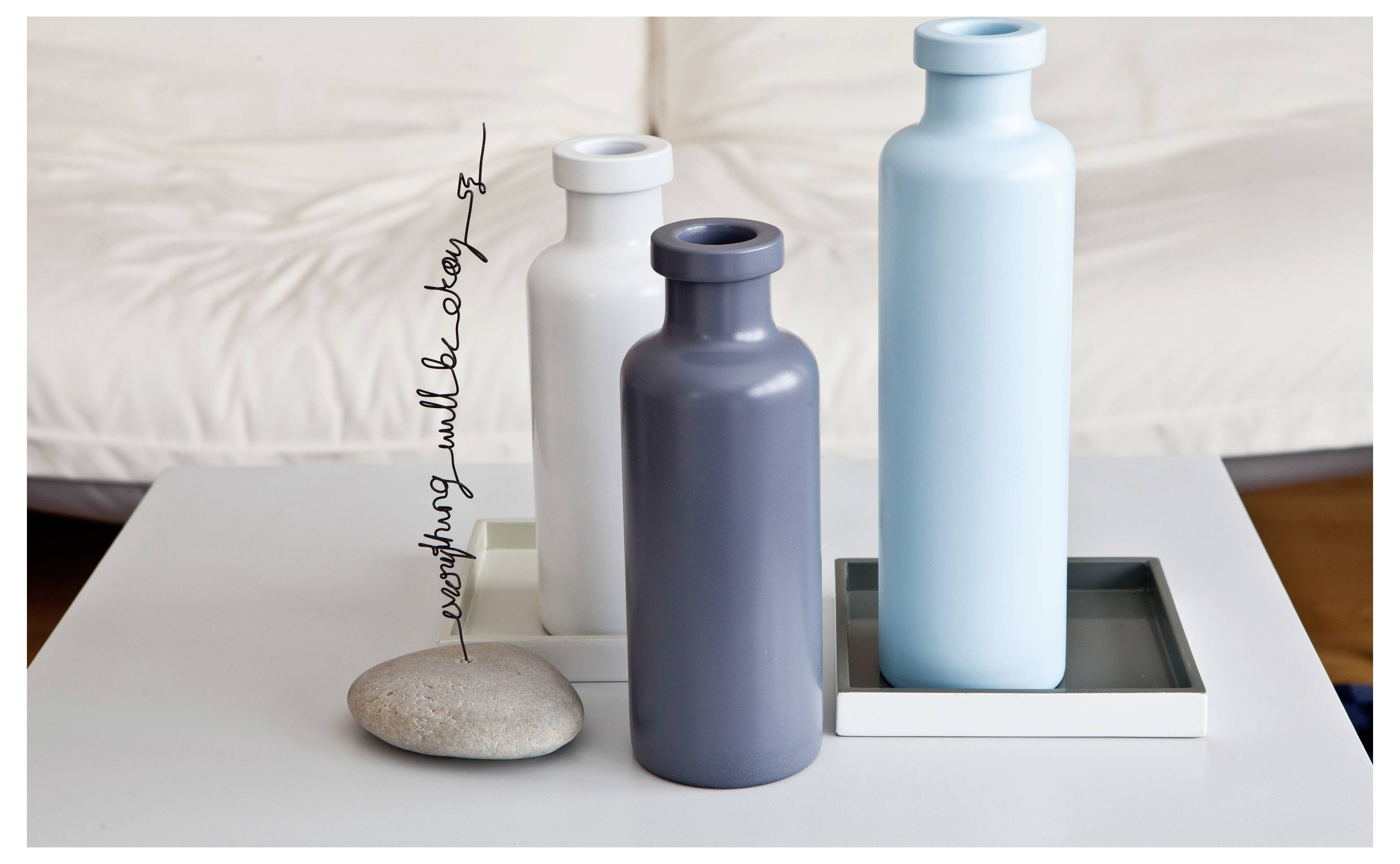 Trois vases en céramique minimaliste sur une table blanche, avec une petite pierre et un texte cursif vertical à côté.