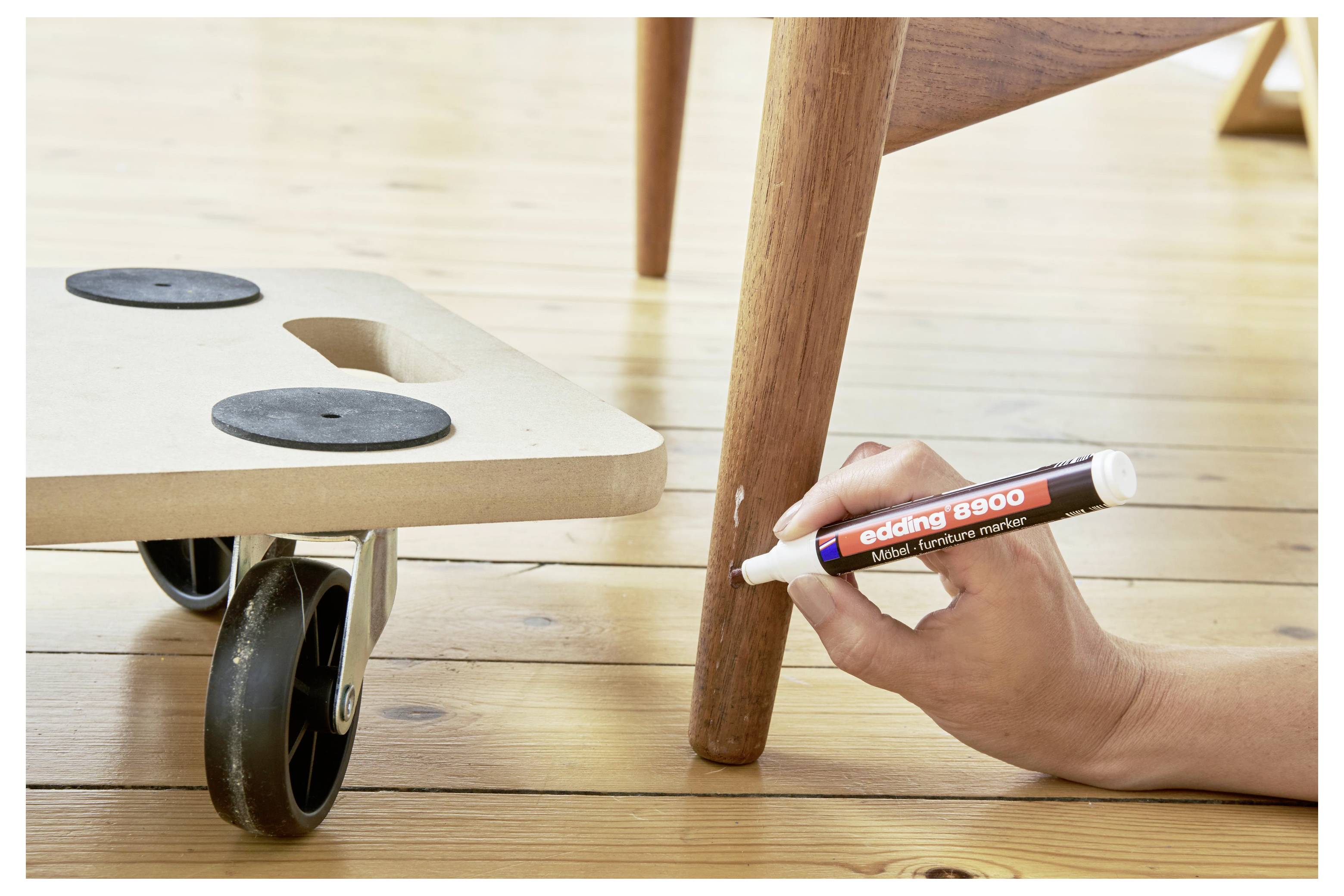 Main utilisant un marqueur pour tracer une ligne sur un pied de chaise en bois, montrant une tâche de réparation de mobilier ou de marquage près d'un siège à roulettes.