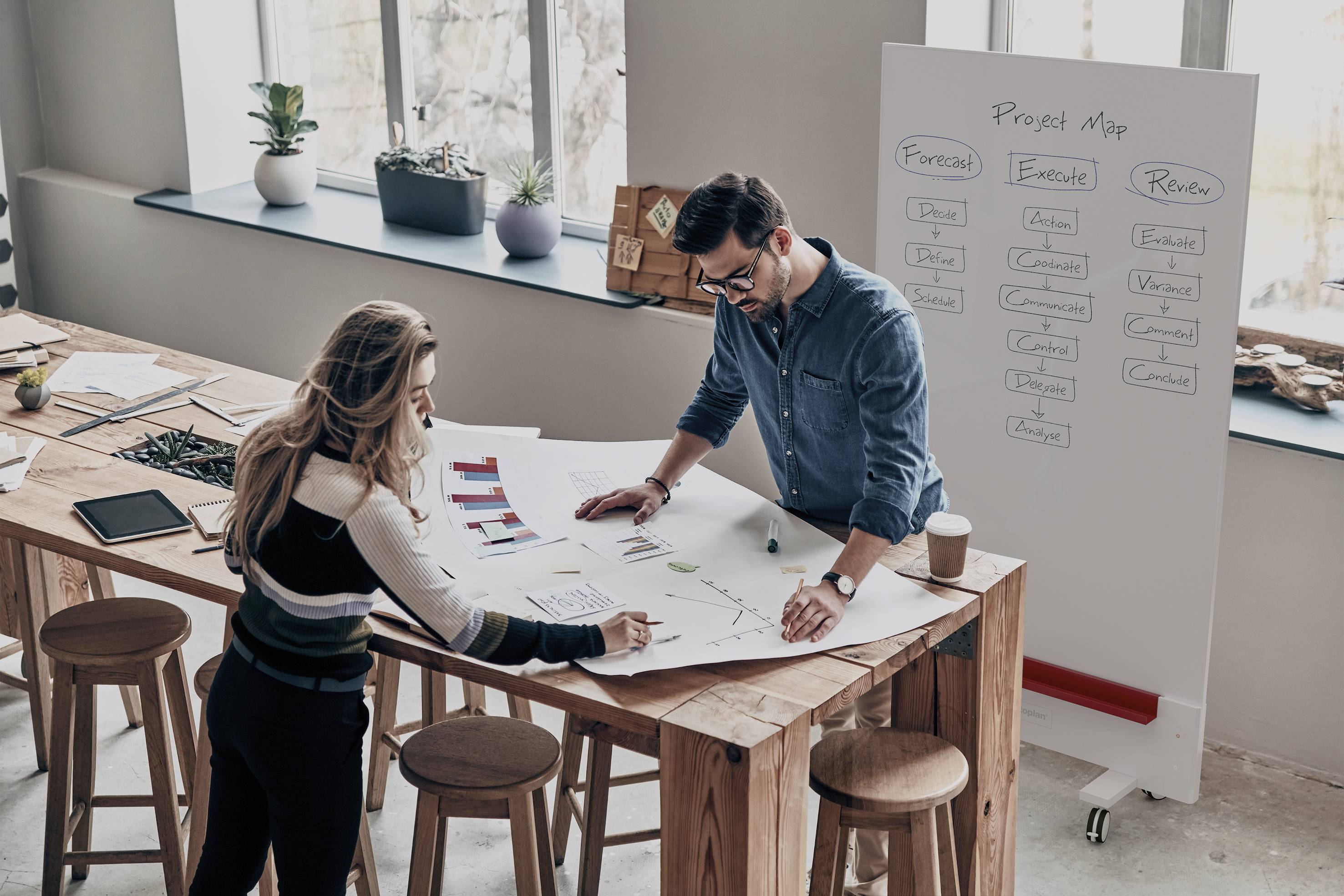 Deux personnes travaillent sur un grand document posé sur une table en bois dans un bureau moderne. Un plan de projet est visible sur un tableau blanc en arrière-plan.