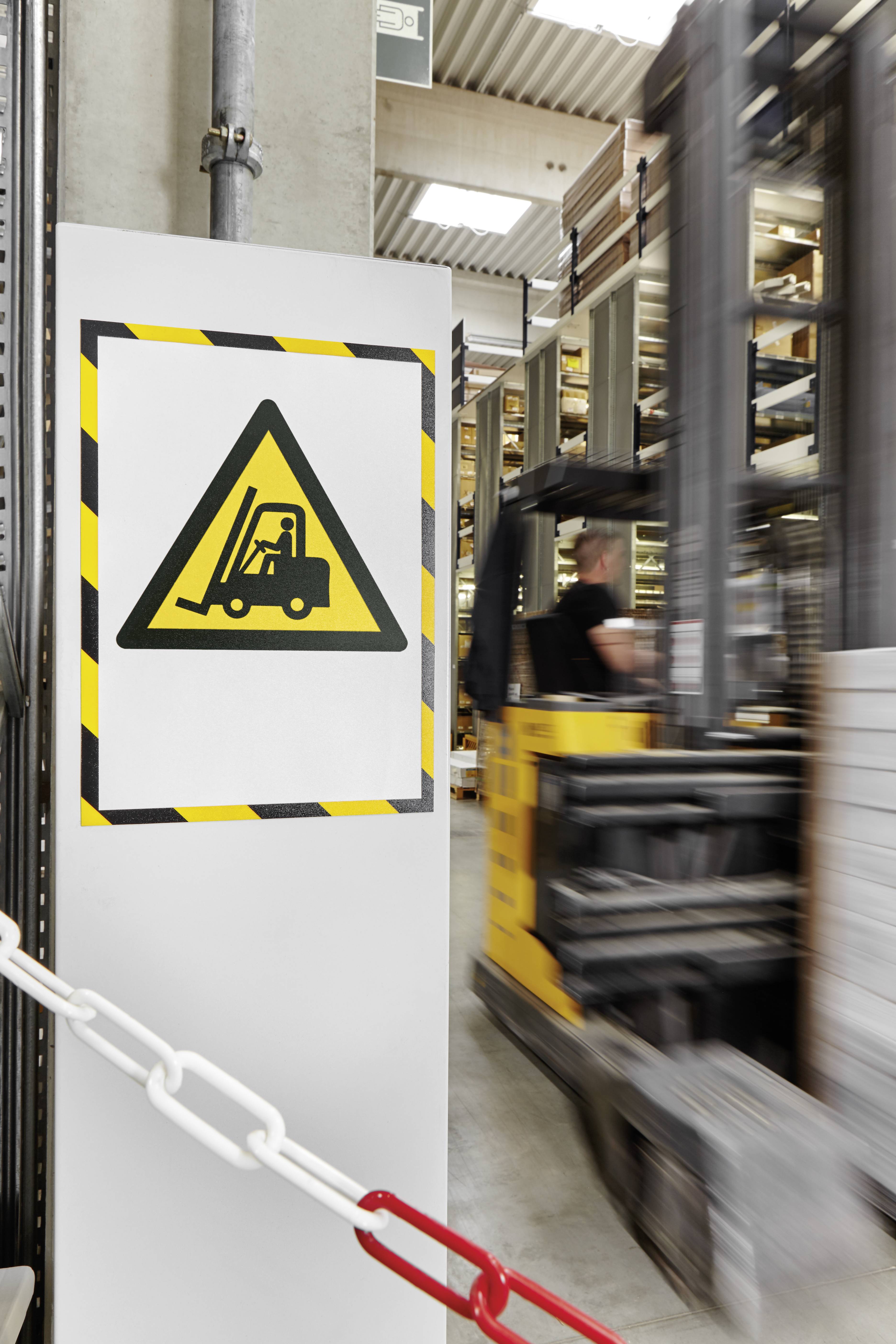Un panneau avec un symbole de chariot élévateur avertit d'une zone de chariots élévateurs dans un bâtiment industriel. À l'arrière-plan, un chariot élévateur en mouvement.