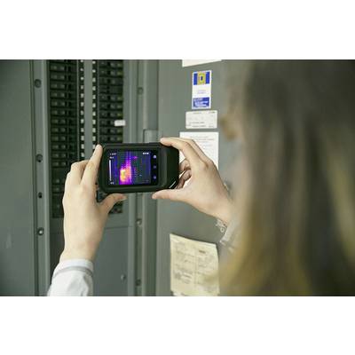 Une personne tient une caméra thermique devant un tableau électrique pour détecter les différences de température. L'écran affiche une thermographie colorée.