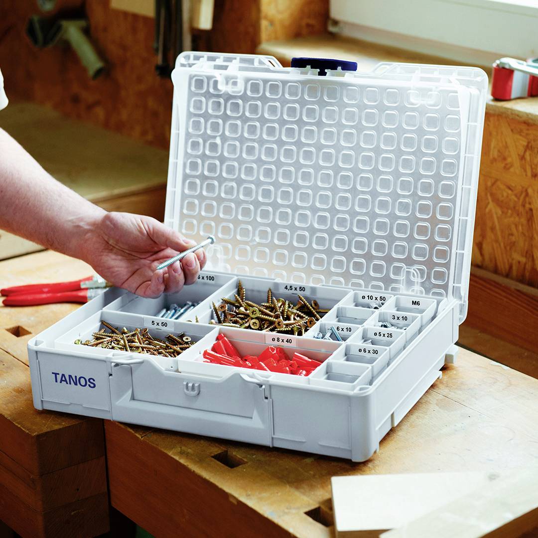 Boîte à outils ouverte sur une table avec des compartiments remplis de vis et de clous. Une main saisit une vis.