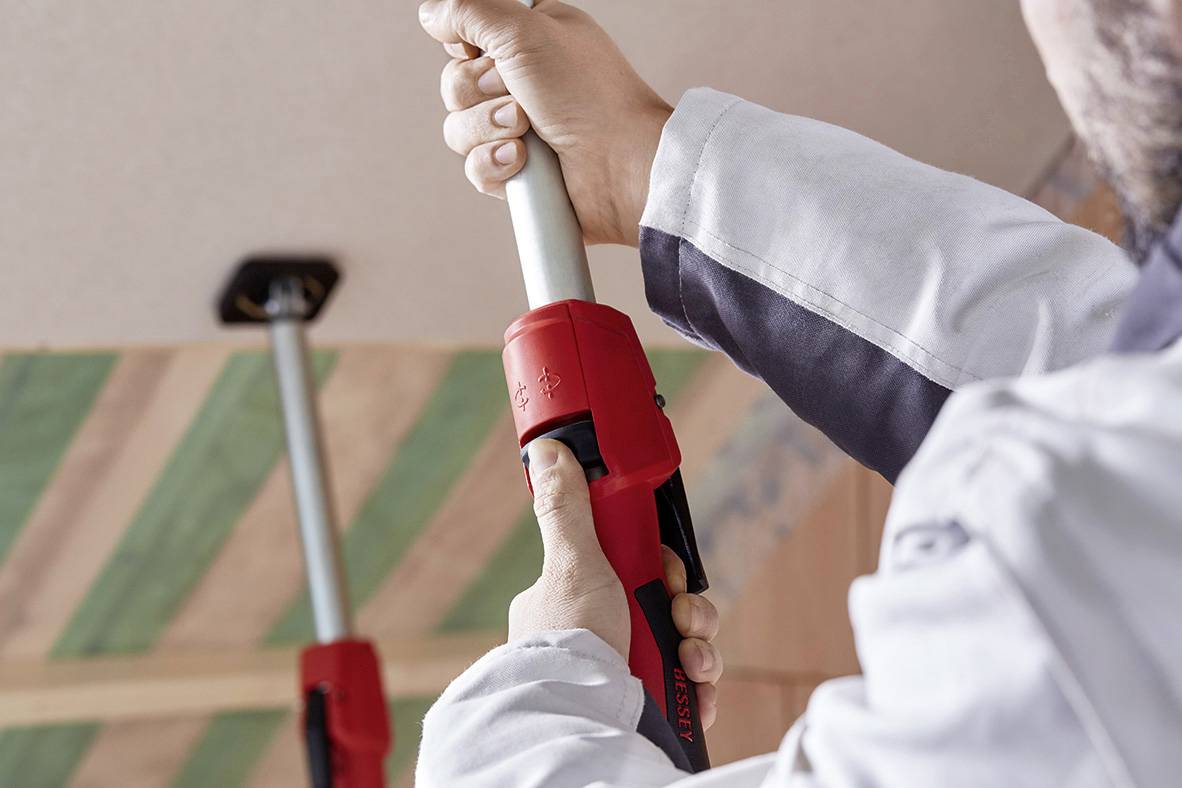 Une personne fixe une perche télescopique au plafond à l'aide d'un système de tension rouge adapté aux travaux de construction.