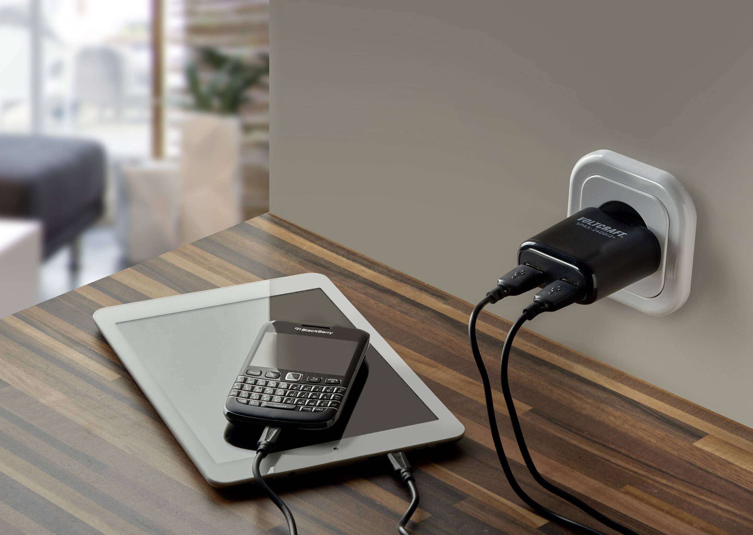 Une tablette et un téléphone mobile sont en train de charger via des câbles de charge branchés à un chargeur USB mural. Les appareils sont posés sur une table en bois.