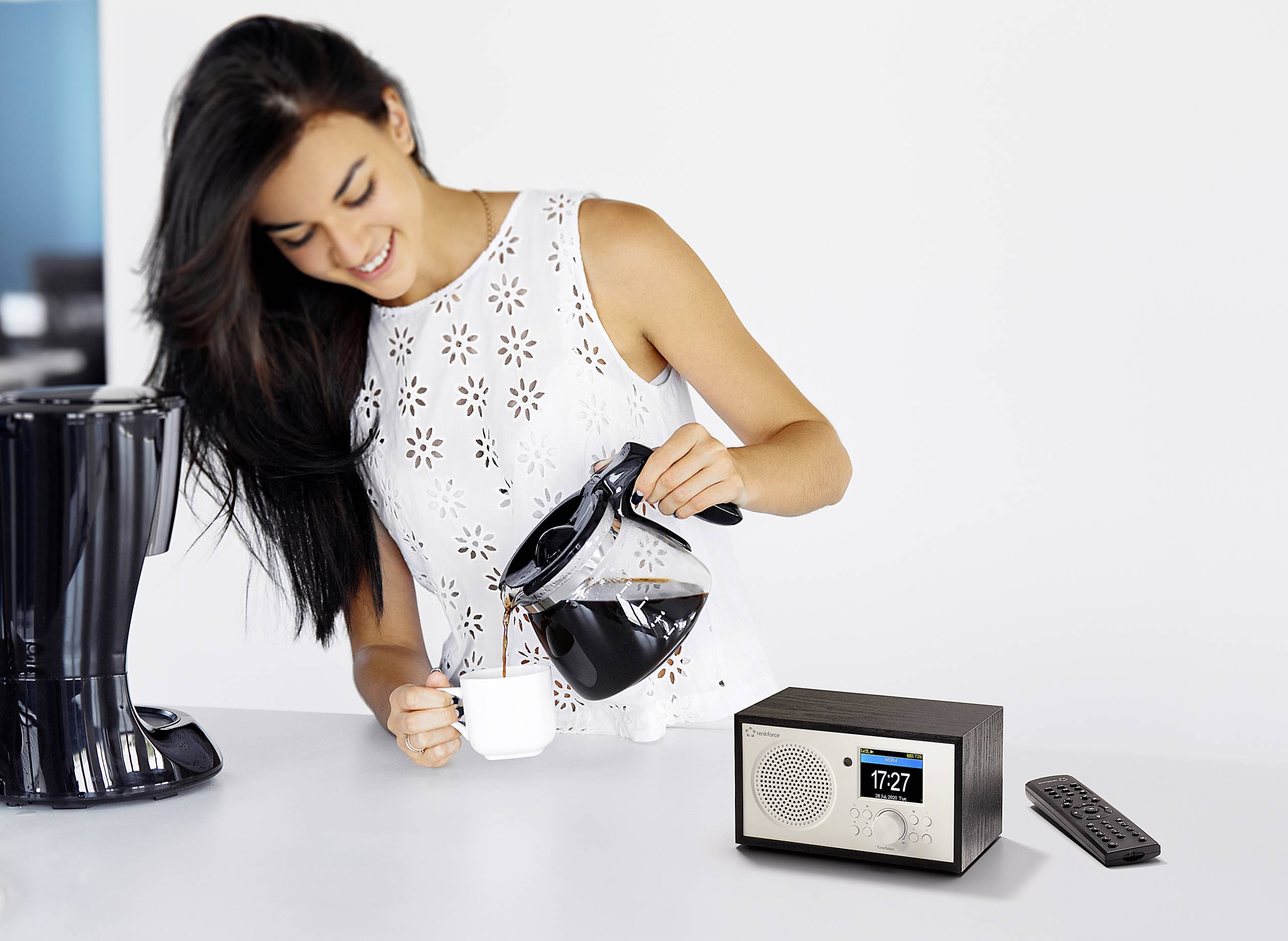 Une femme verse du café dans une tasse à côté d'une radio numérique affichant l'heure '17:27' sur une table.