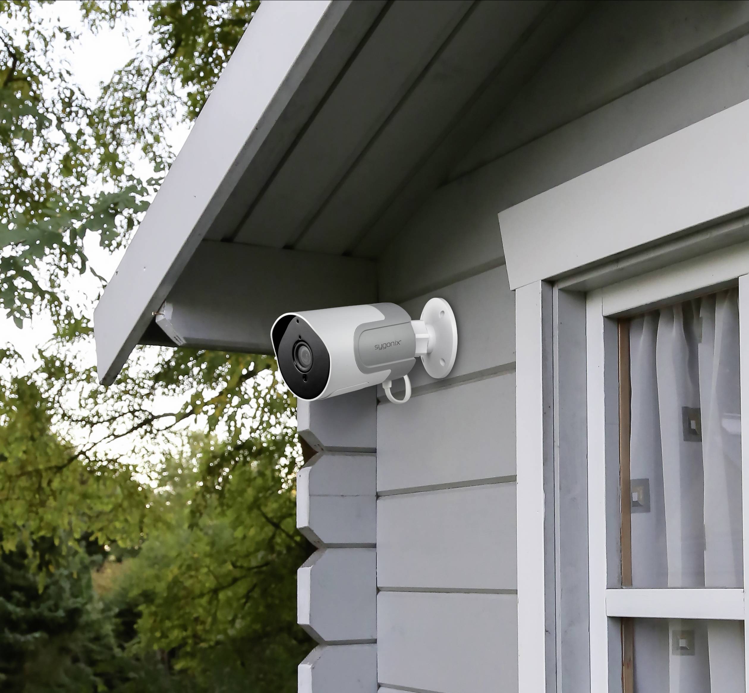 Caméra de surveillance montée sur le mur extérieur d'une maison, à côté d'une fenêtre ; des arbres visibles en arrière-plan.