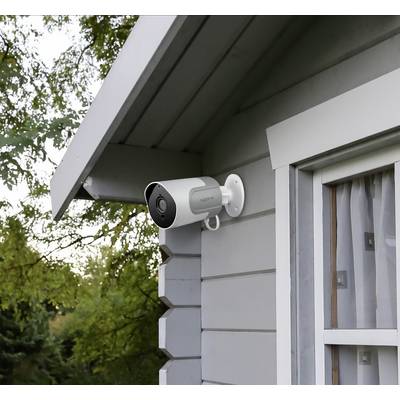 Caméra de surveillance montée sur le mur extérieur d'une maison, à côté d'une fenêtre ; des arbres visibles en arrière-plan.