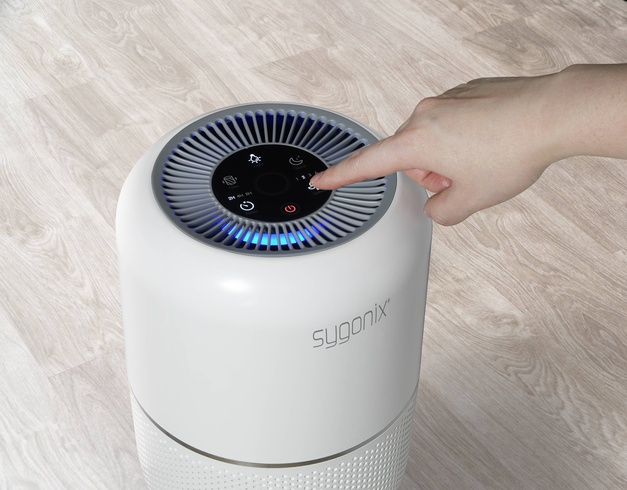 Une main touche le dessus d'un purificateur d'air rond blanc sur un sol en bois. L'appareil dispose d'un panneau de commande éclairé.