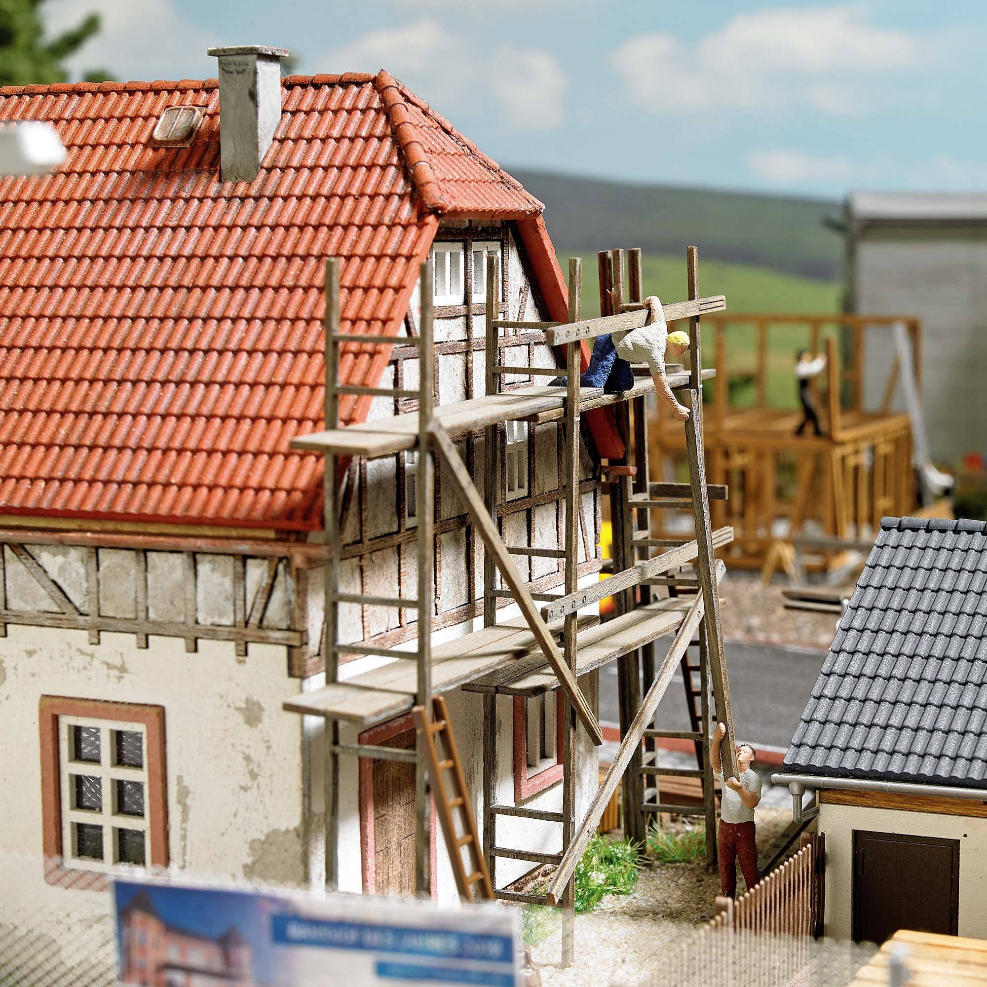 Une petite maquette d'une maison à colombages en construction, avec des échafaudages. À l'arrière-plan, d'autres bâtiments et un pré vert.