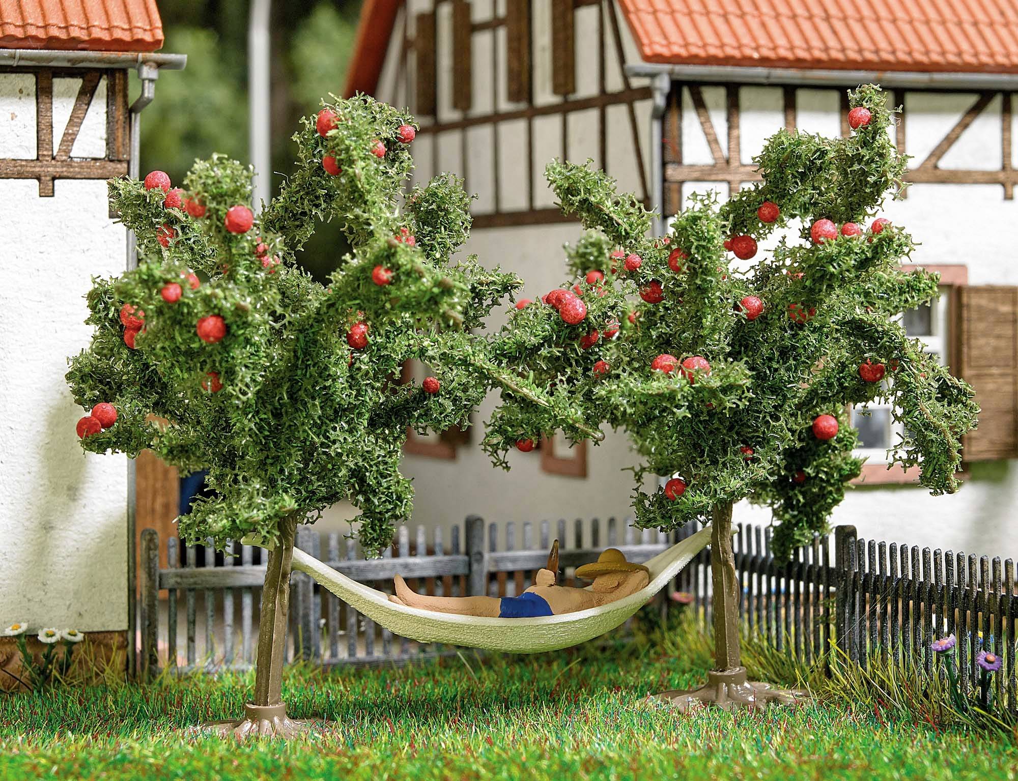 Un hamac est suspendu entre deux pommiers, où une personne est allongée. Les arbres portent de nombreuses pommes rouges. En arrière-plan, on aperçoit des maisons à colombages.