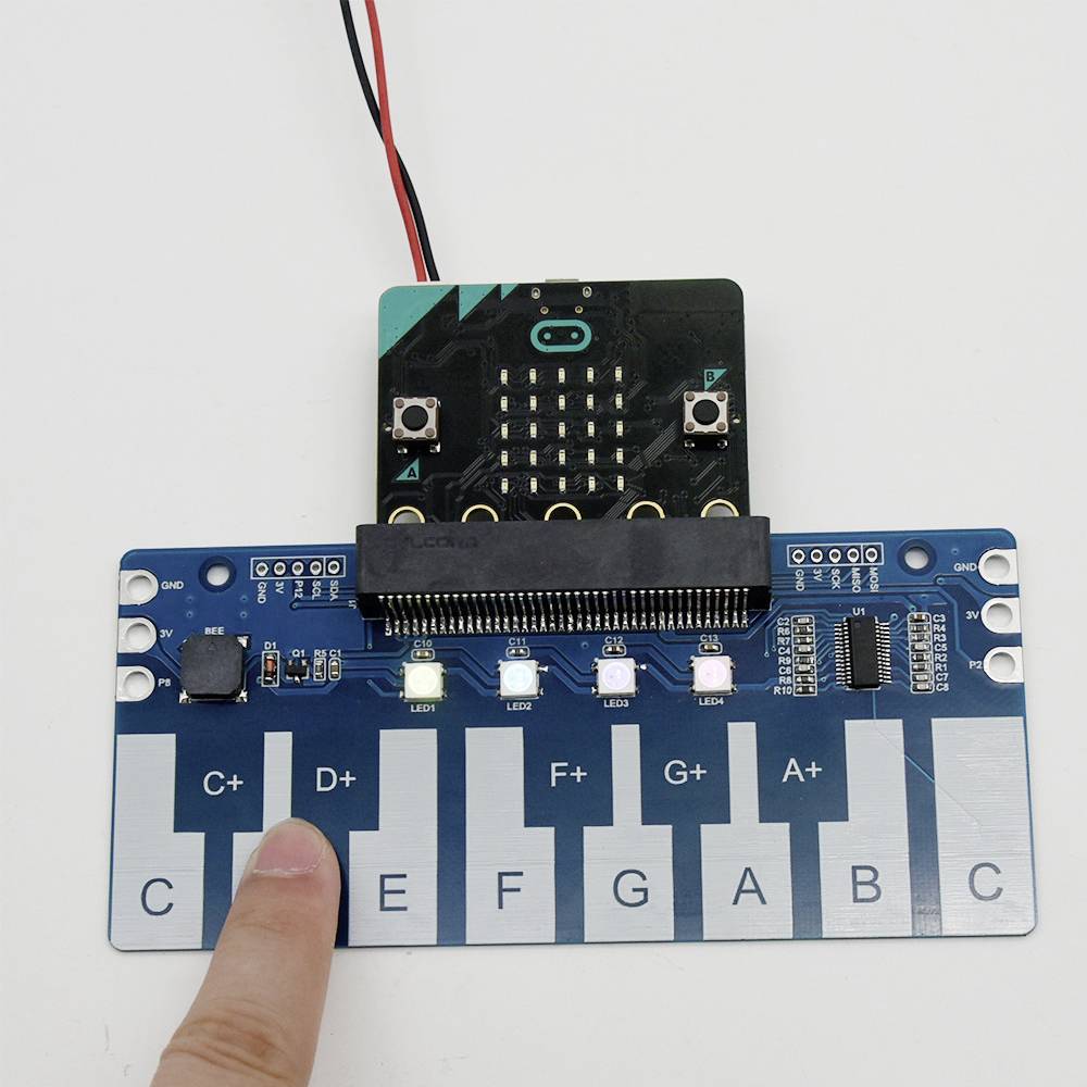 Un doigt pointe vers un module de piano électronique équipé d'un microcontrôleur. Le module possède six touches représentant des notes.