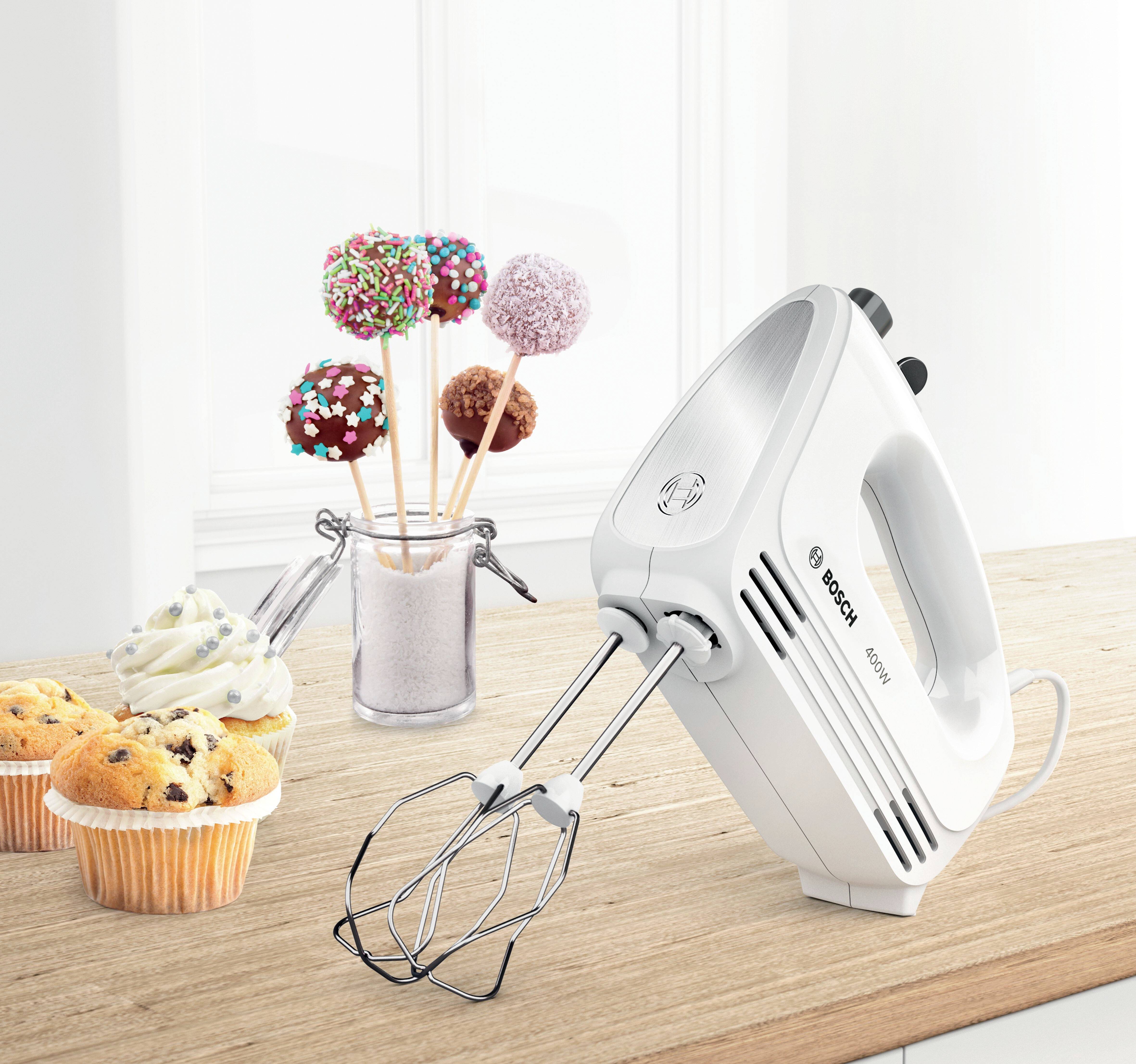Un batteur électrique blanc est posé sur un plan de travail de cuisine à côté de sucettes décoratives et de muffins garnis de crème.