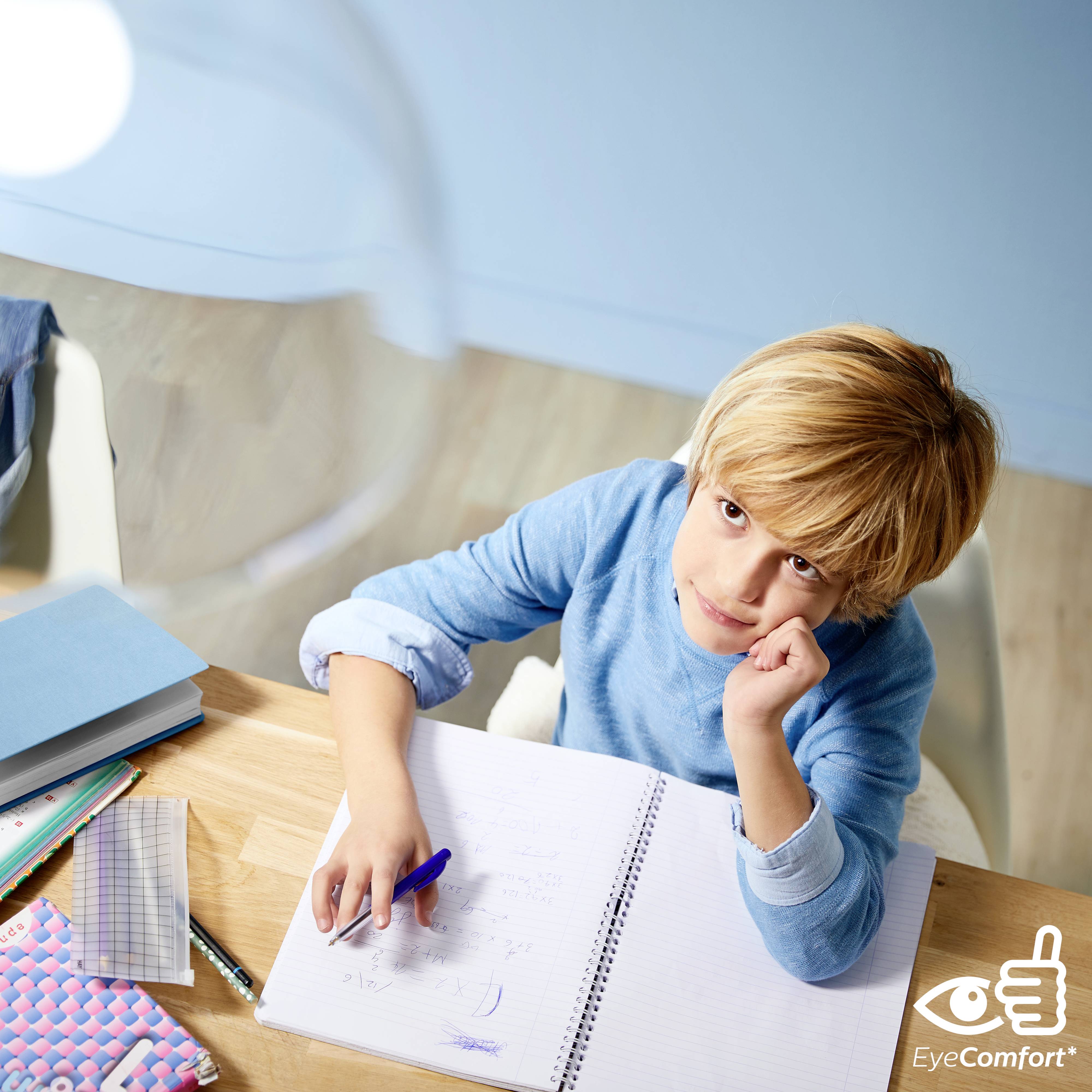 Un enfant est assis à une table, regard pensif, et écrit dans un cahier. Du matériel scolaire est éparpillé sur la table.