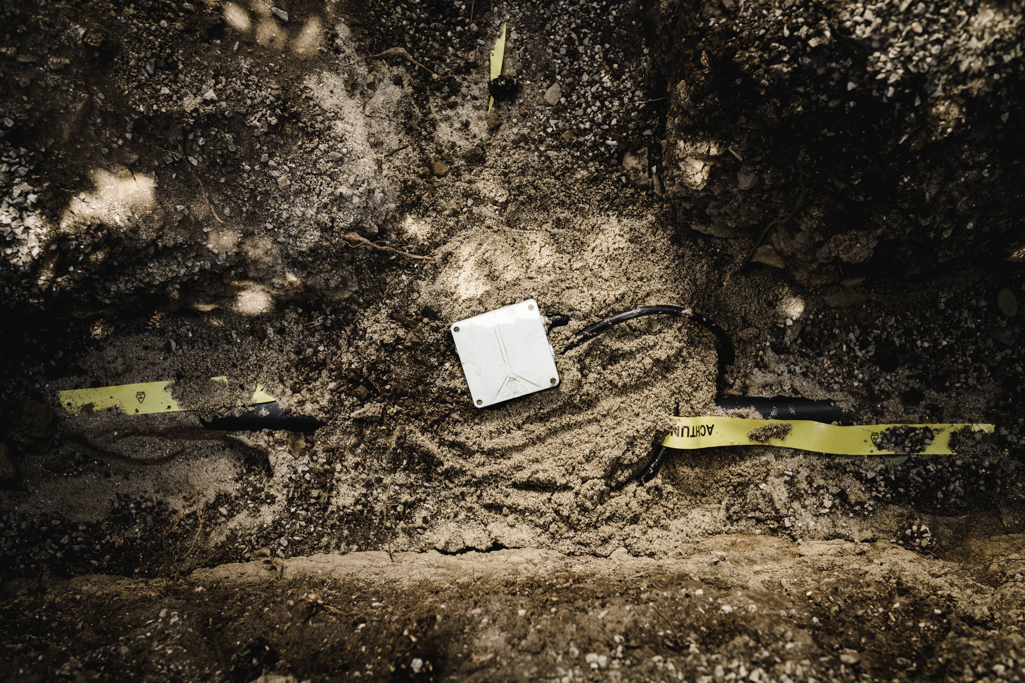 Une boîte de jonction électrique enterrée dans un sol sablonneux, entourée d'un ruban de signalisation jaune.