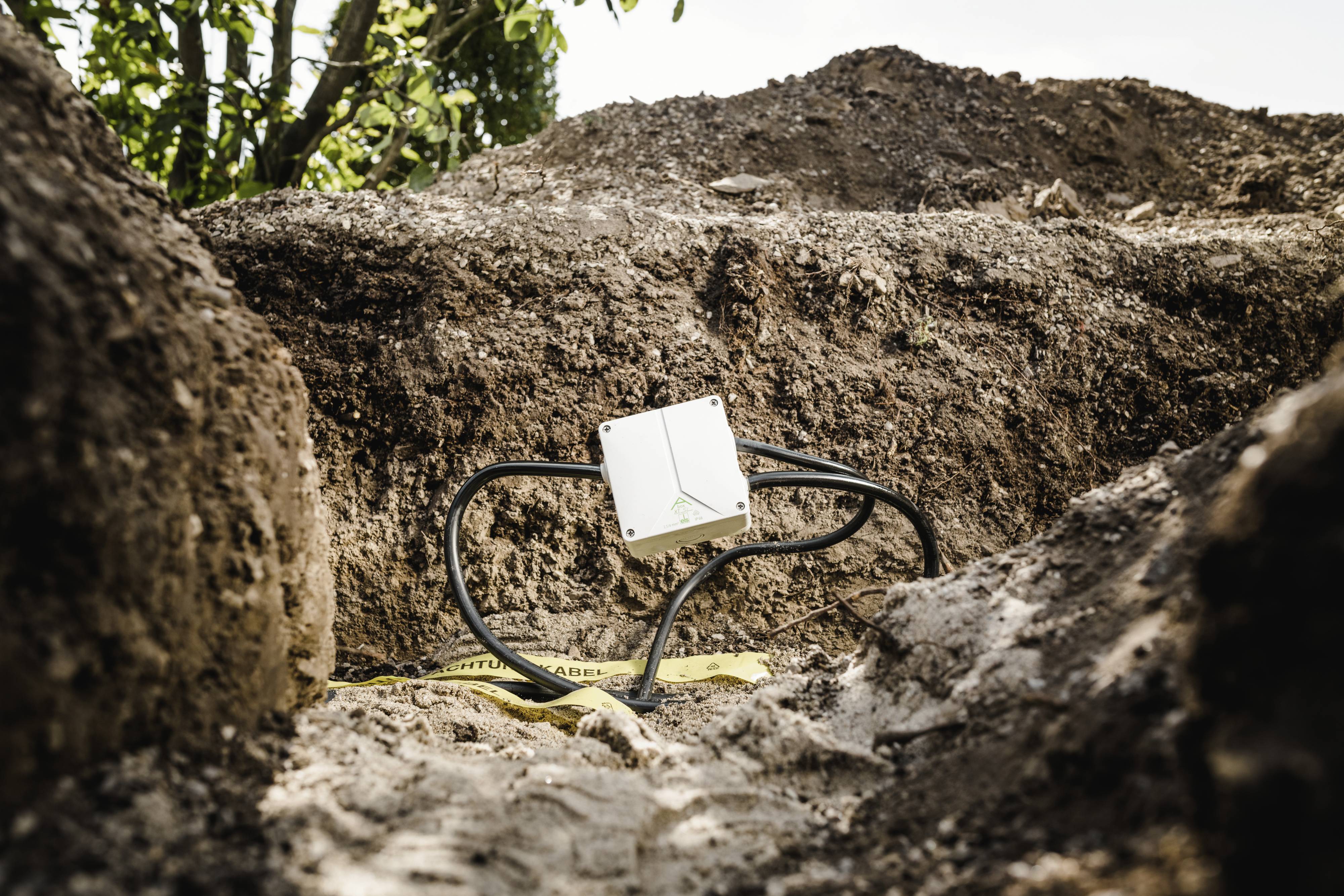 Un petit appareil blanc avec des câbles est posé dans une tranchée. L'arrière-plan montre de la terre et des plantes, probablement lors de travaux archéologiques.