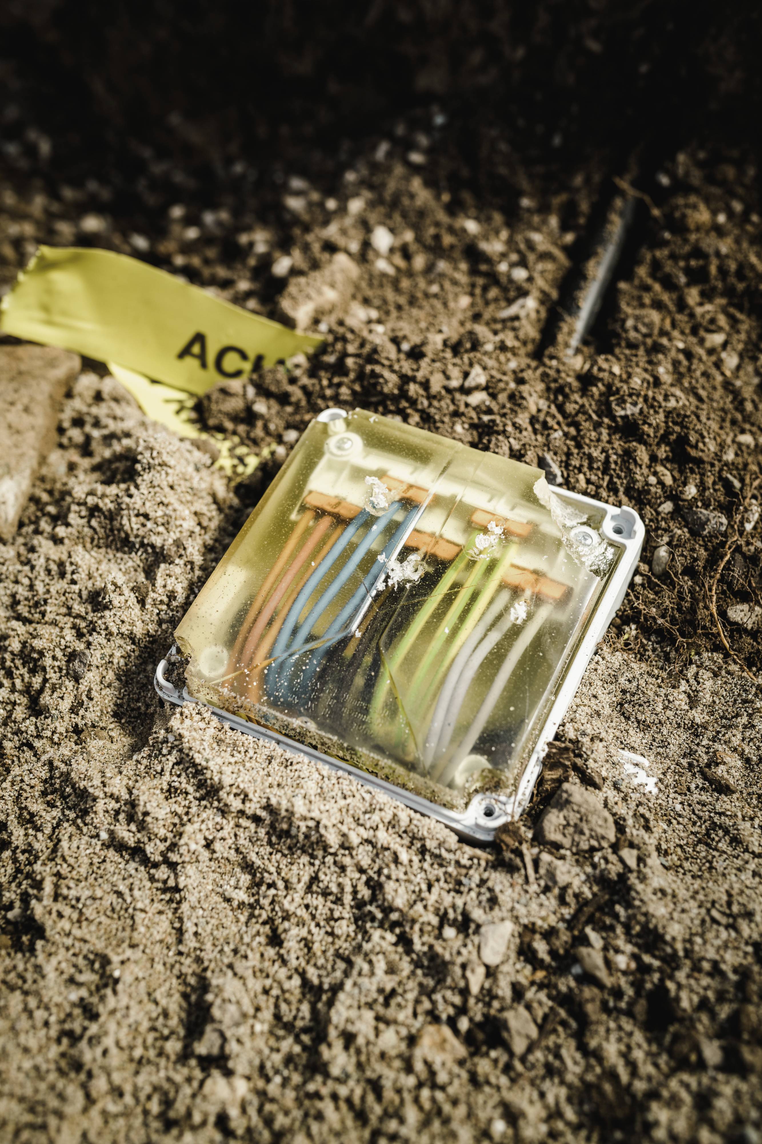 Une prise ouverte avec des fils de couleur dans le sol révèle des câbles exposés, entourés de saleté et de ruban de signalisation jaune.