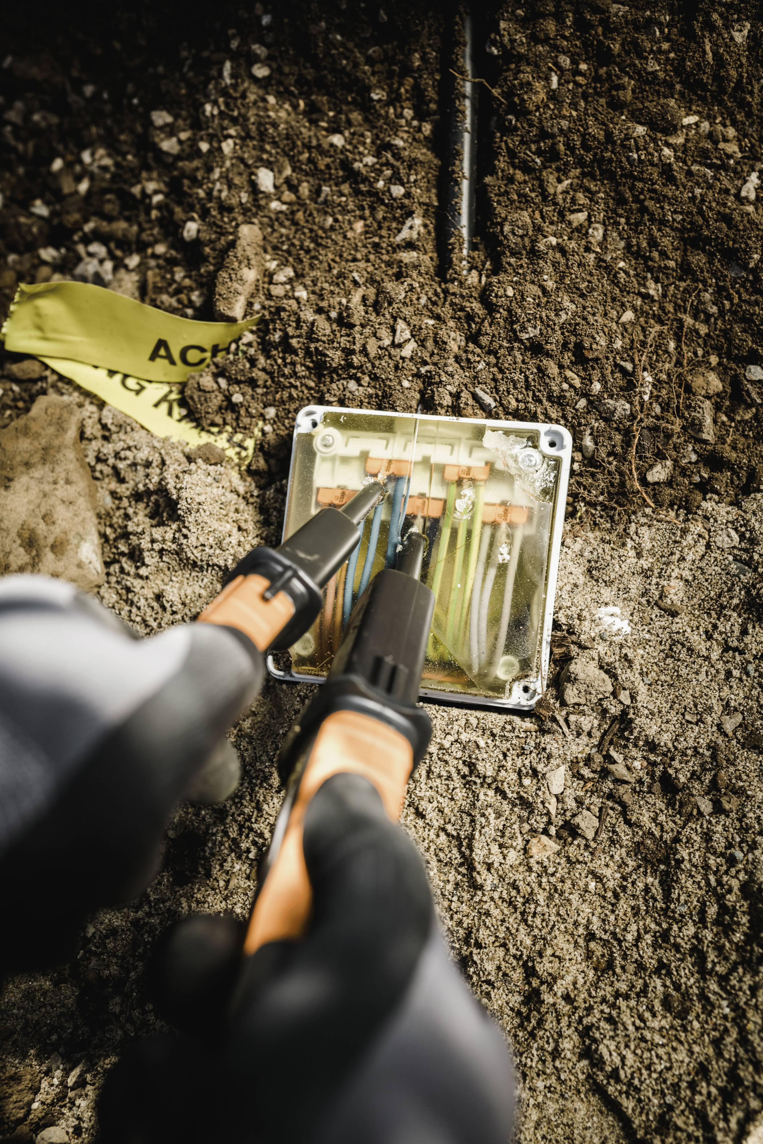 Un électricien travaille à l'extérieur sur un boîtier de distribution électrique ouvert, connectant plusieurs fils de différentes couleurs. Un ruban de signalisation jaune est visible en arrière-plan.