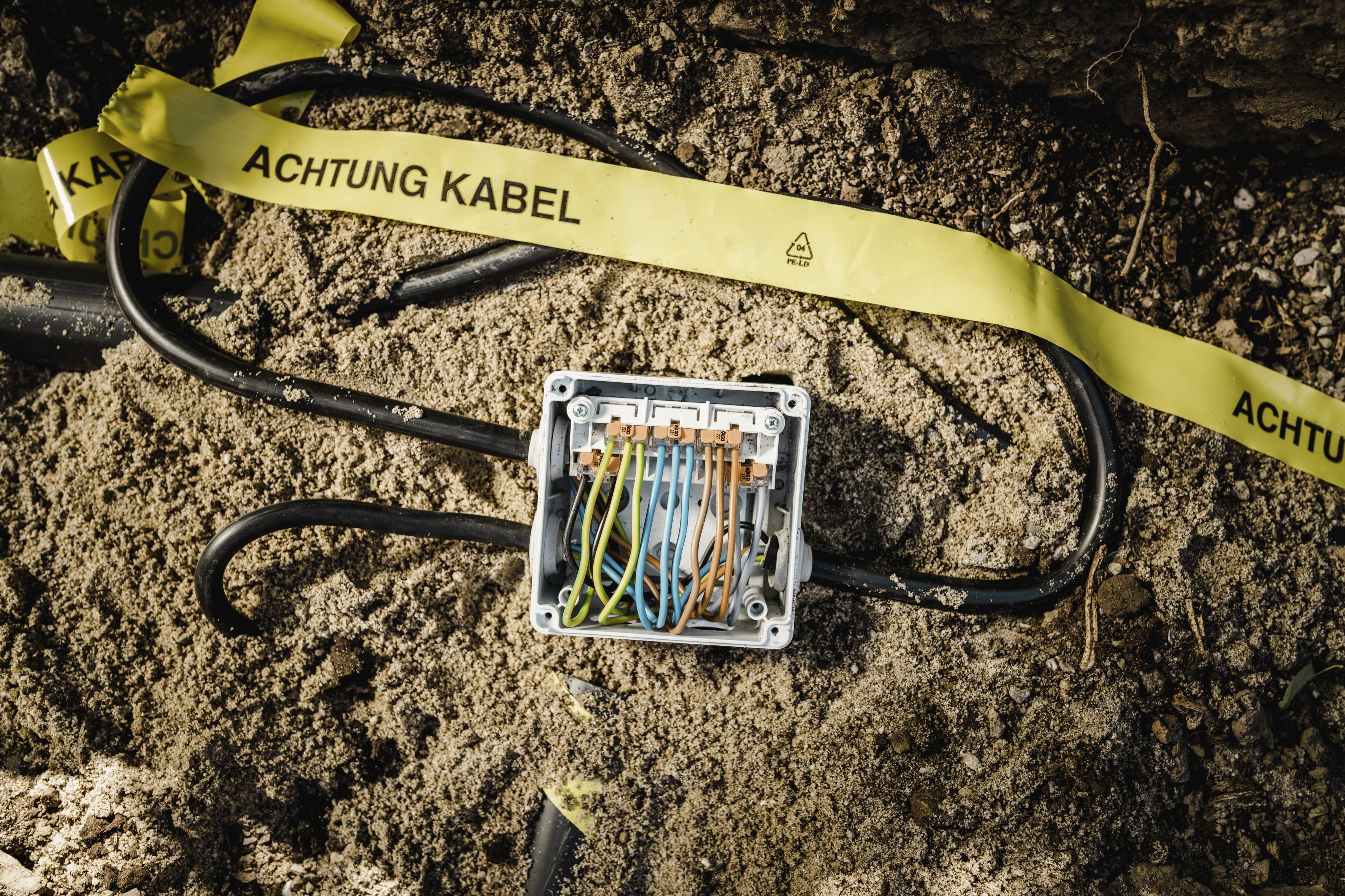 Une boîte de jonction ouverte avec des câbles colorés dans le sol. Un ruban jaune au-dessus portant l'inscription « Attention câbles », pour signaler des travaux sur les câbles.