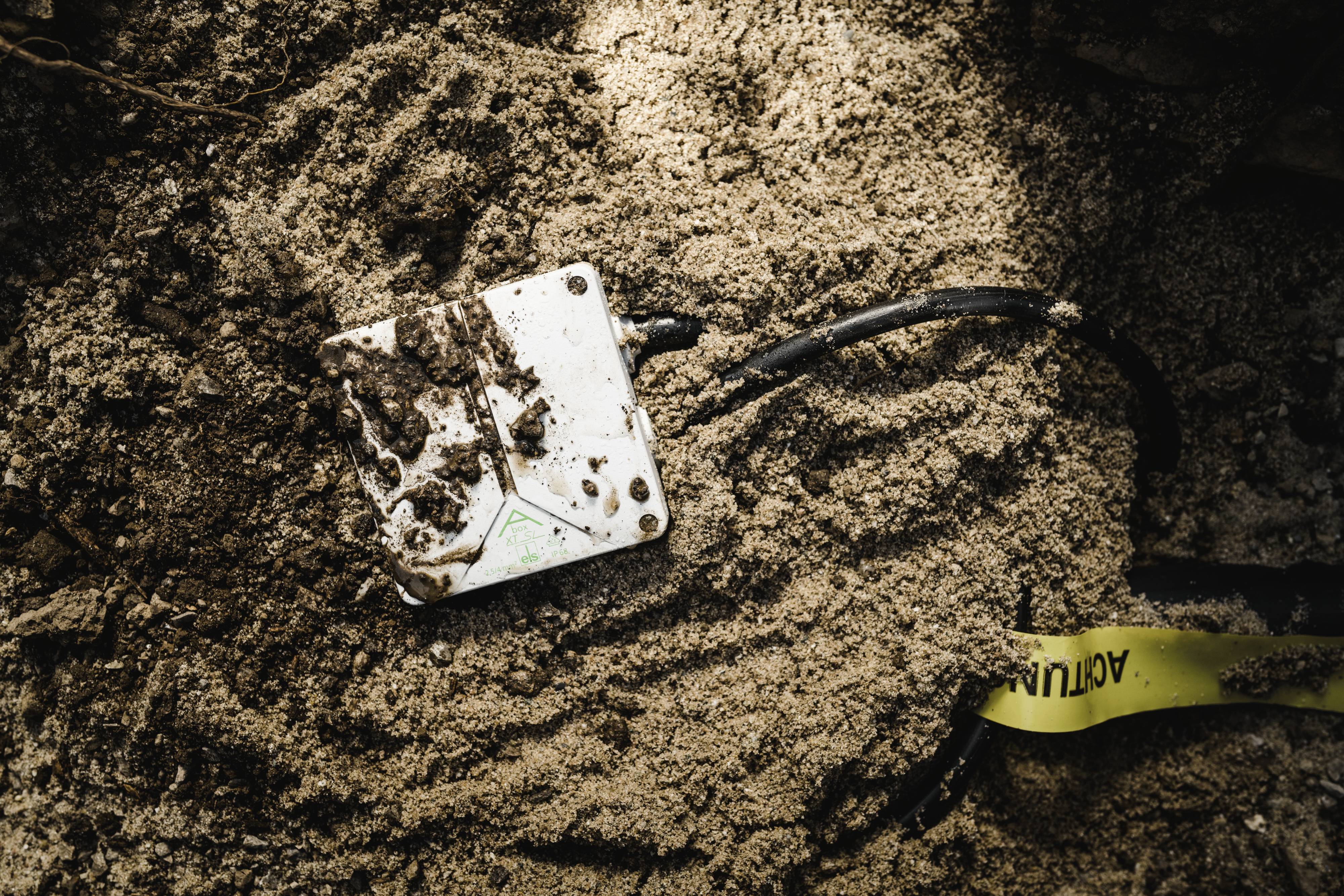 Un appareil blanc sale avec des câbles est posé sur un sol sablonneux, partiellement recouvert de saleté.