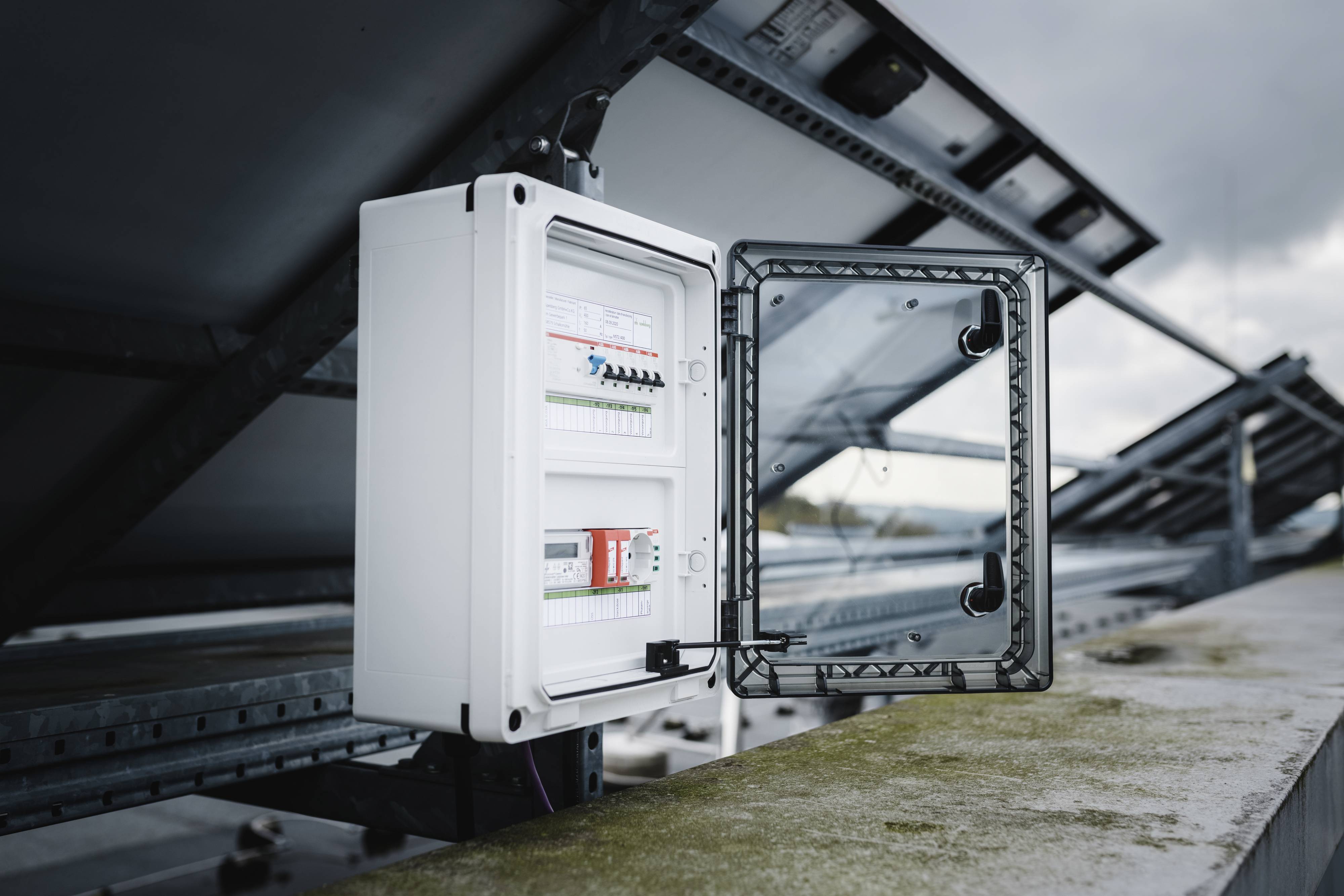 Un tableau électrique ouvert sur un toit avec des modules solaires installés en arrière-plan, sous un ciel nuageux.