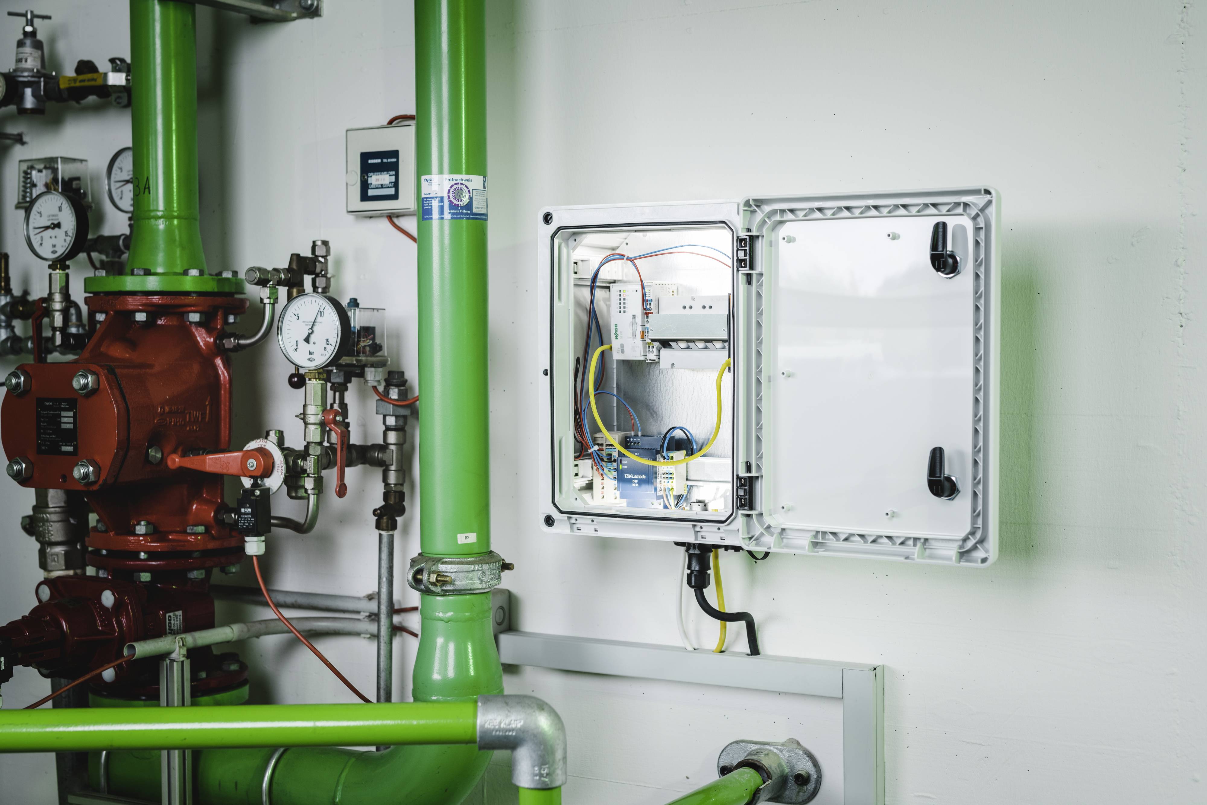 Armoire de distribution ouverte sur un mur blanc, entourée de tuyaux verts et de manomètres. Le boîtier révèle le câblage électrique et ses composants.
