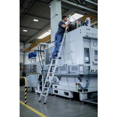 Un homme est debout sur une échelle et travaille sur une grande machine grise dans un environnement industriel. La machine porte des étiquettes et des circuits électriques.