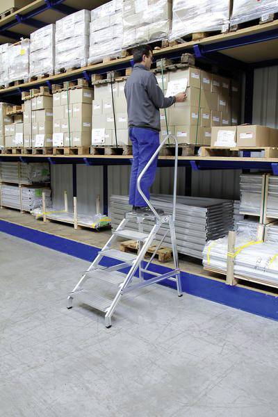 Une personne est debout sur une échelle métallique et vérifie des cartons dans une étagère d'entrepôt. Les étagères sont remplies de différentes caisses et paquets.