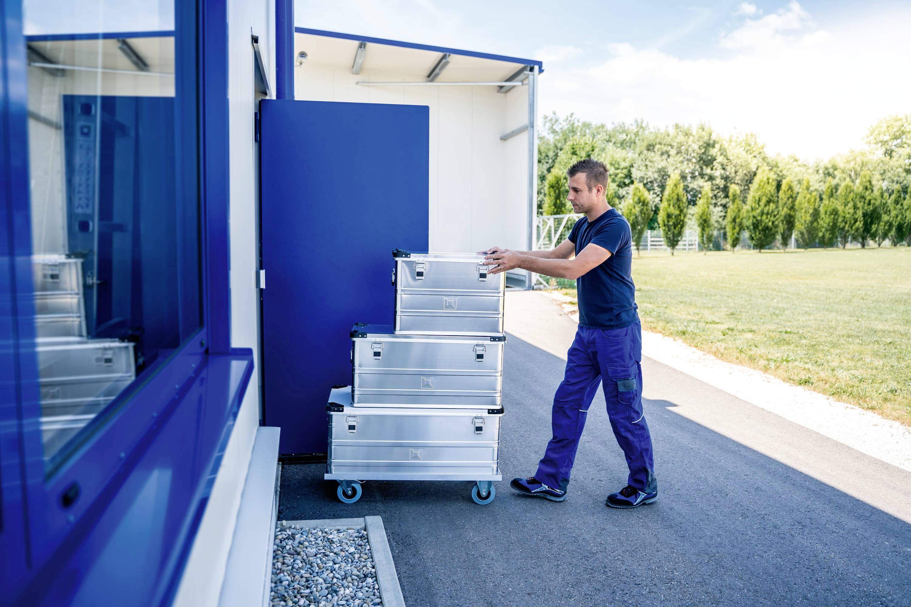 Un homme pousse une pile de caisses argentées sur un chariot roulant vers un bâtiment. Des arbres et un pré sont visibles en arrière-plan.