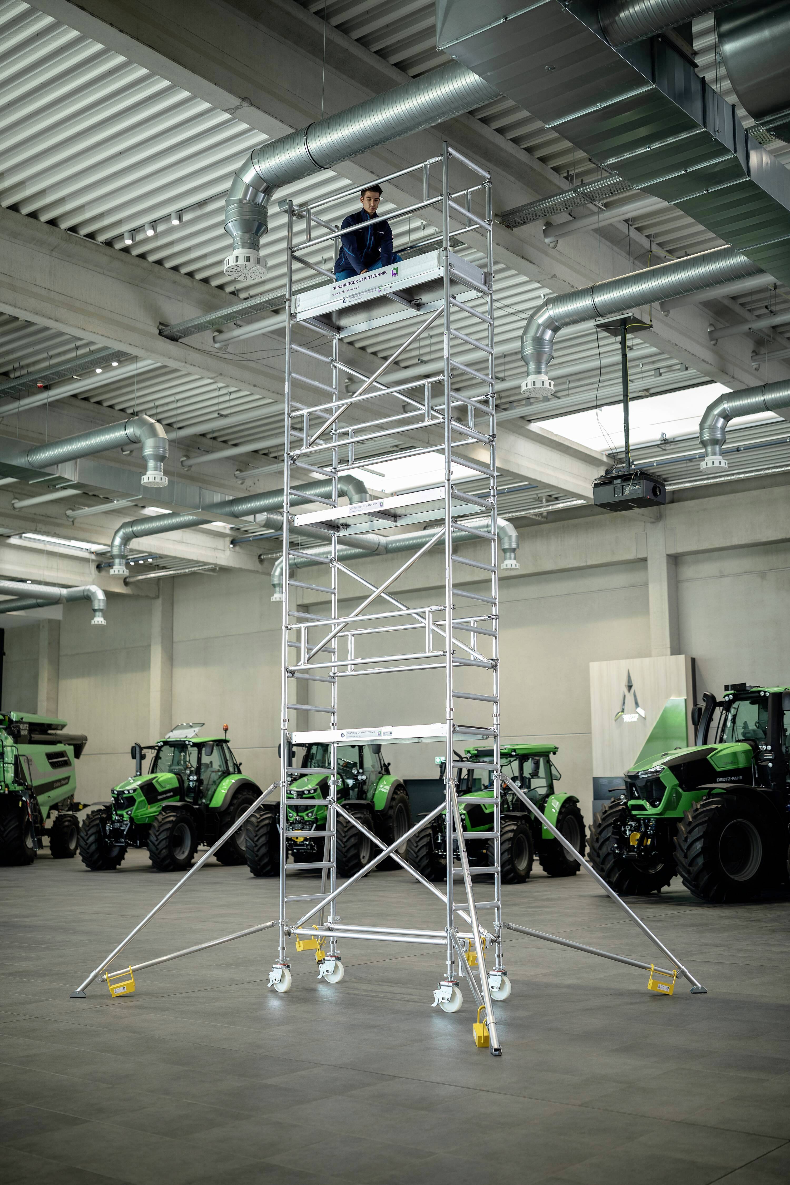 Une personne se tient sur un échafaudage mobile élevé dans un grand hall, avec des tracteurs verts en arrière-plan, prête à procéder à une inspection.