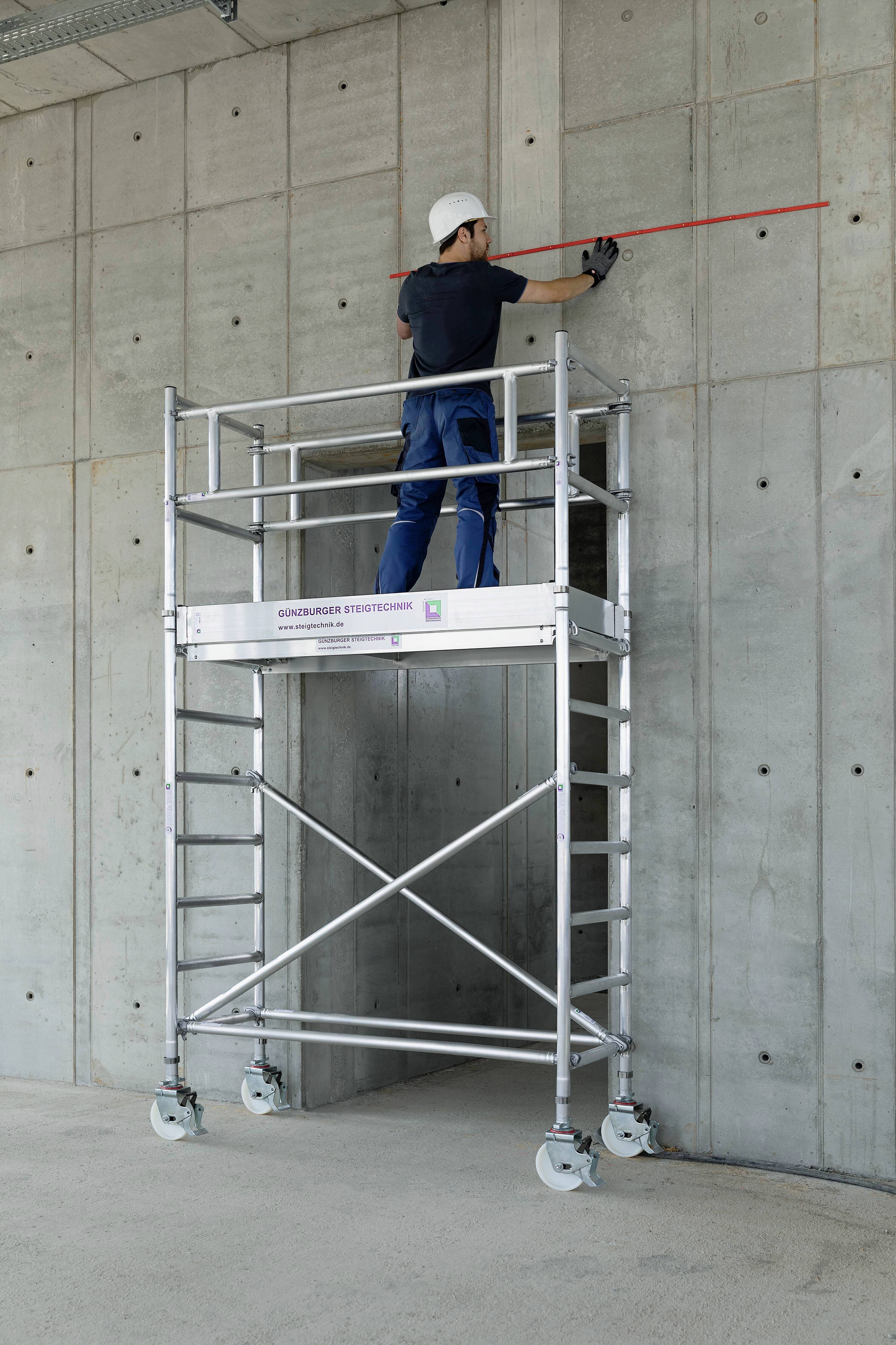 Un ouvrier portant un casque se tient sur un échafaudage et mesure un mur en béton avec une tige rouge.