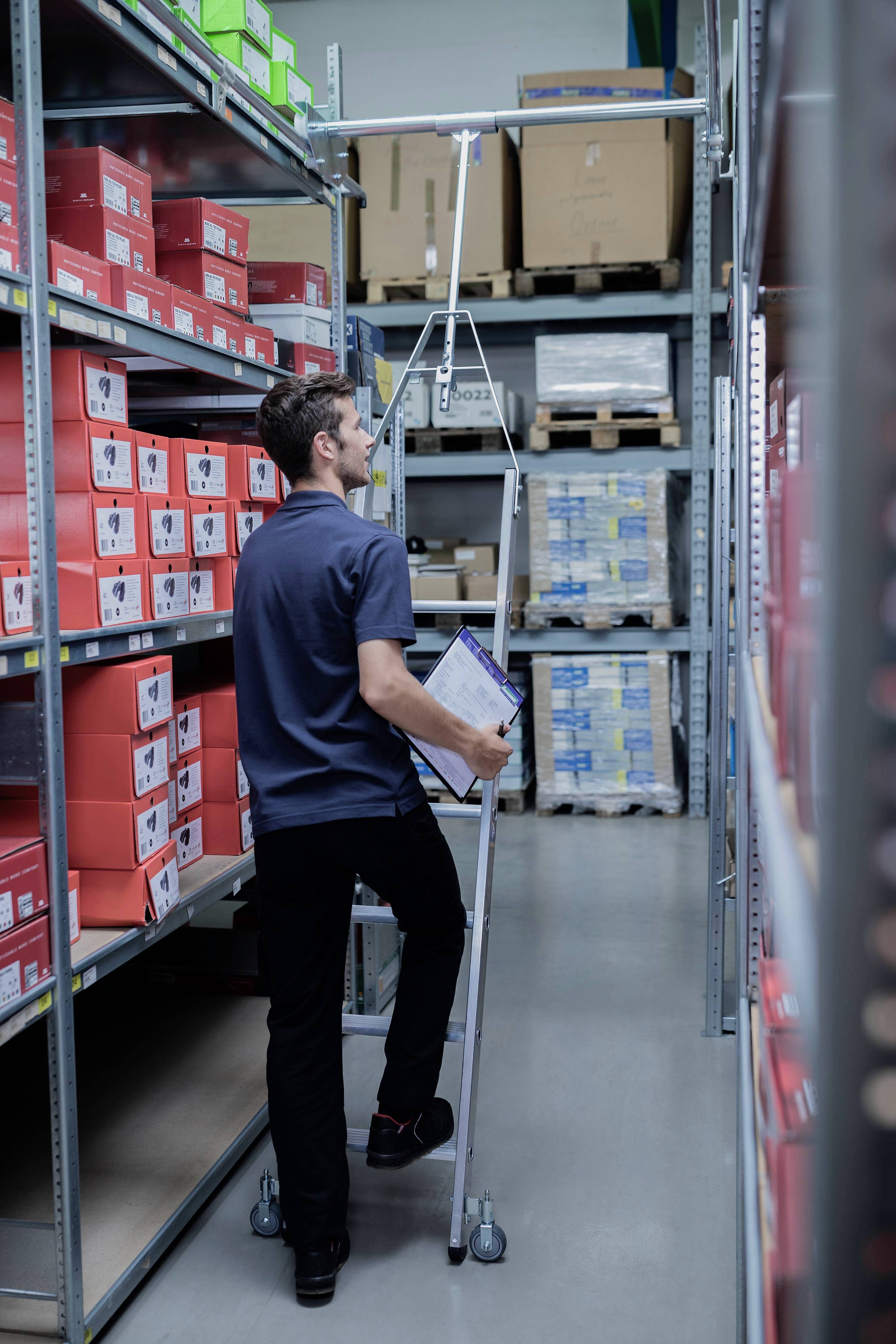 Un homme dans un entrepôt soulève un carton d'une étagère haute à l'aide d'un bras télescopique. Il tient un presse-papiers dans son autre main.