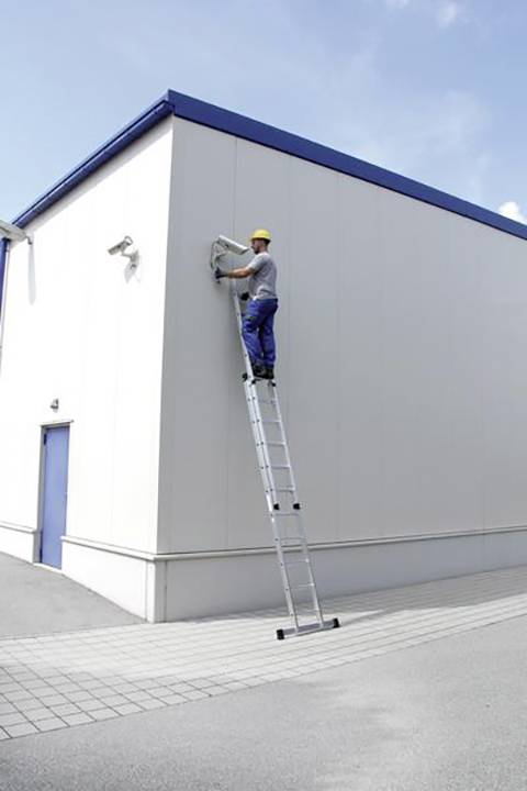 Un travailleur sur une échelle répare un climatiseur sur un bâtiment. Casque et équipement de protection présents. Conditions météorologiques dégagées.