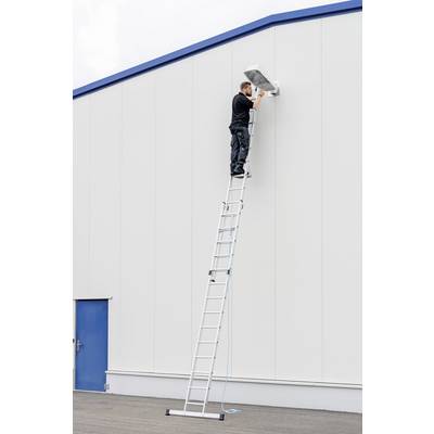 Une personne se tient au sommet d'une longue échelle, en train d'installer une caméra de surveillance sur un mur latéral élevé d'un bâtiment.
