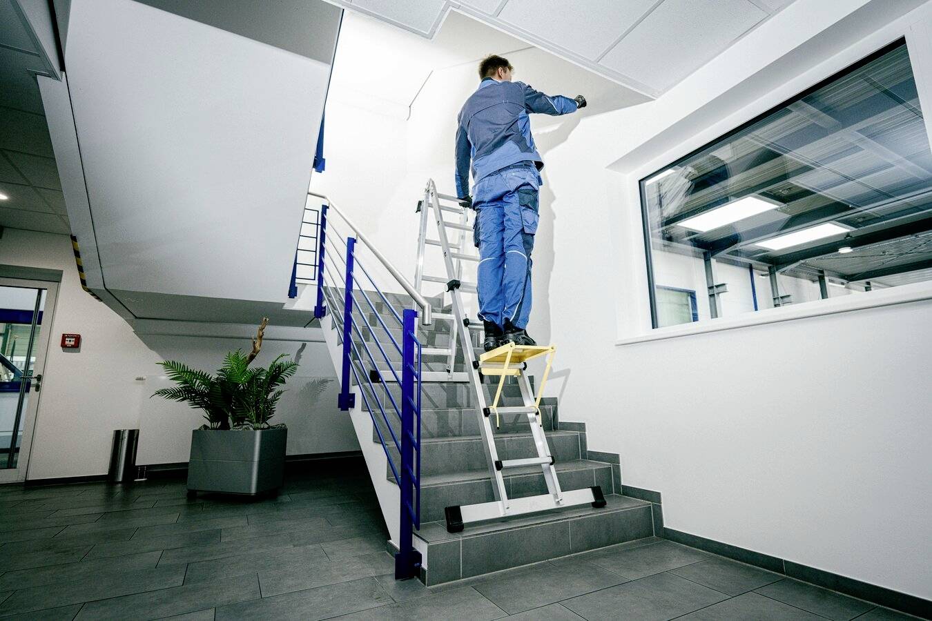Un ouvrier sur une échelle peint le plafond d'un escalier dans un immeuble de bureaux moderne. Il est bien équipé et porte des vêtements de travail.