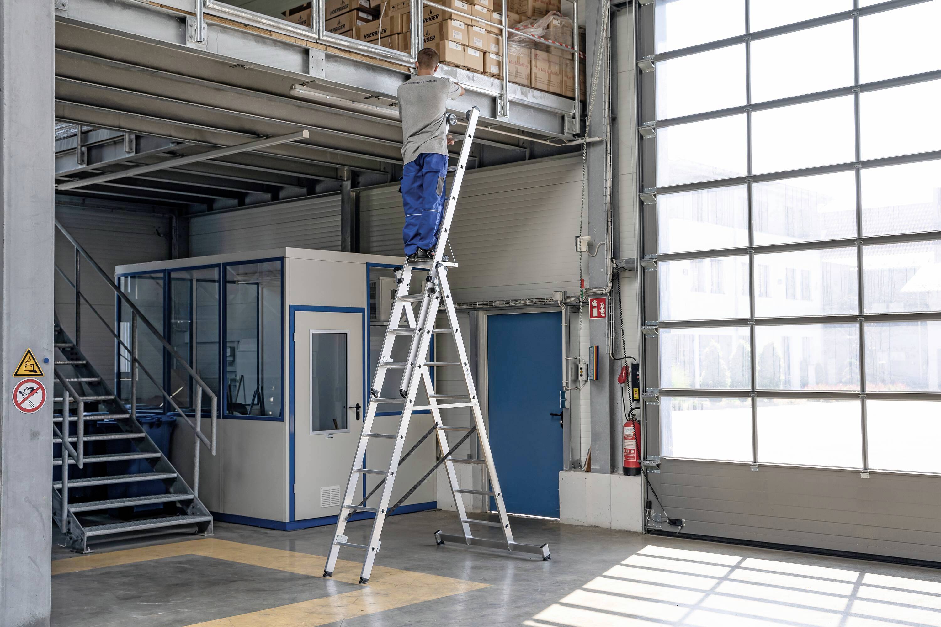 Une personne est debout sur une échelle dans un atelier, travaillant sur une étagère. Une porte bleue et une cabine vitrée sont visibles en arrière-plan. La sécurité est primordiale.