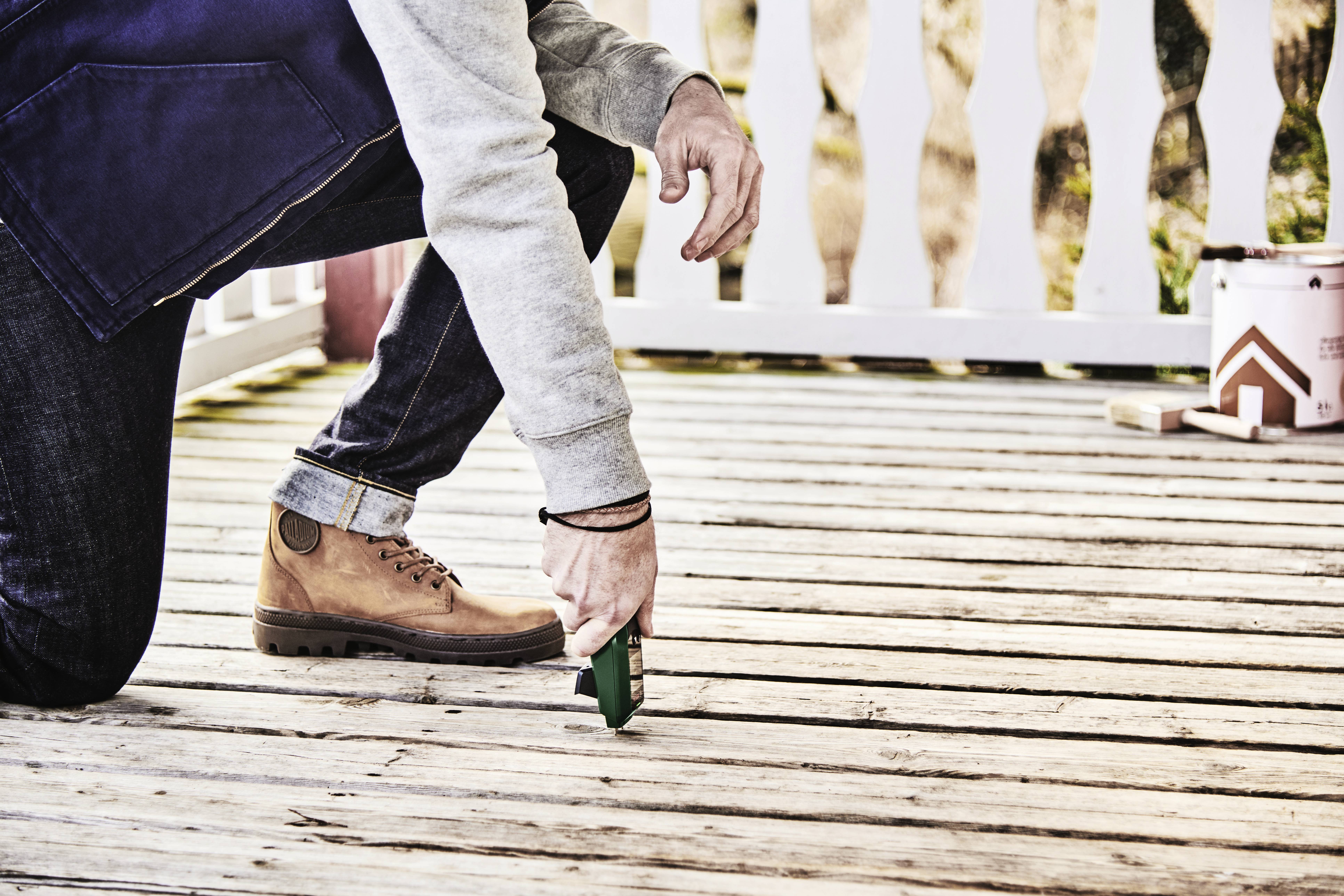 Une personne est agenouillée et construit une terrasse en utilisant un outil. Un clôture blanche est visible en arrière-plan.