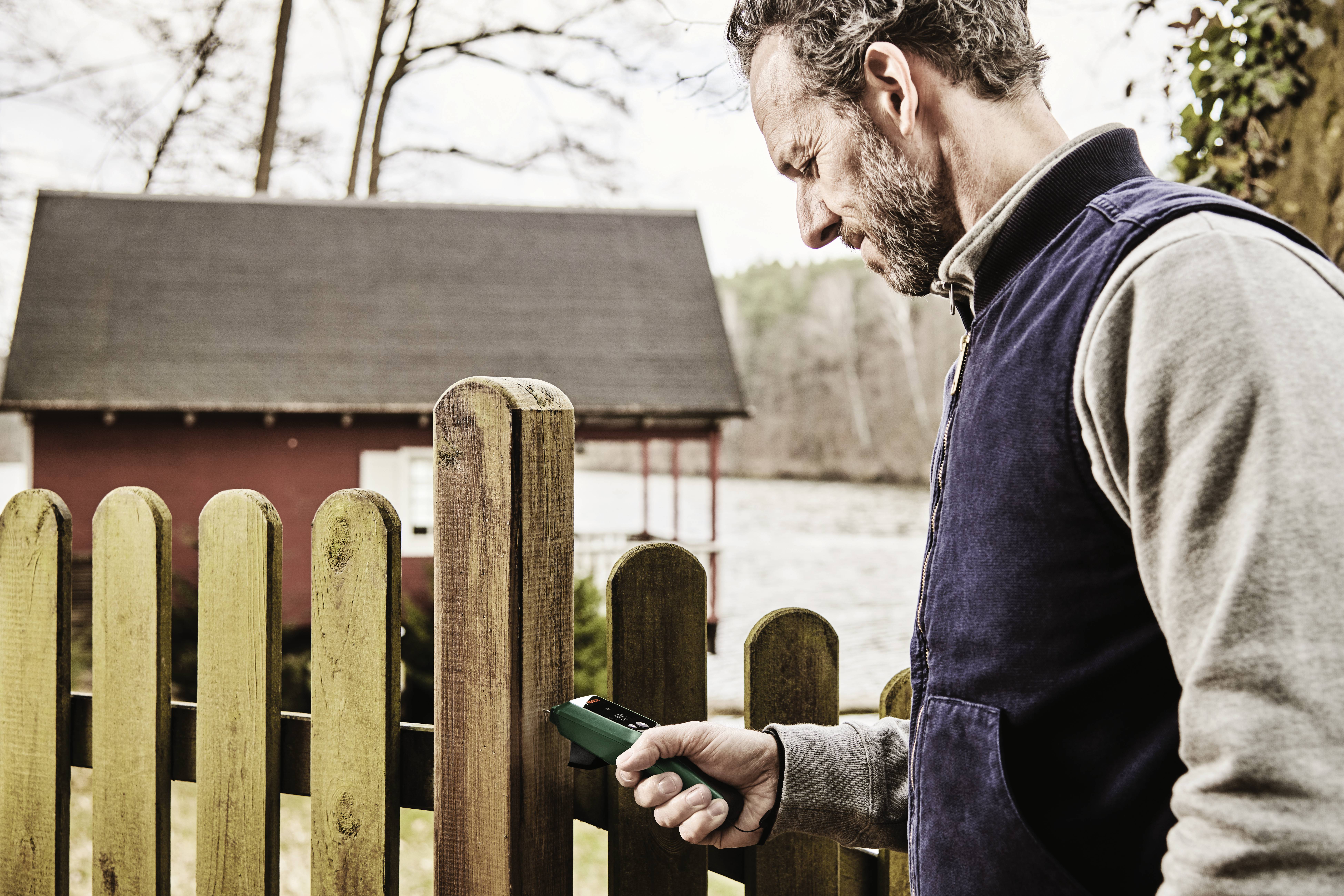 Un homme mesure l'humidité d'une clôture en bois à l'aide d'un appareil. À l'arrière-plan, on aperçoit une maison située au bord d'une rivière.