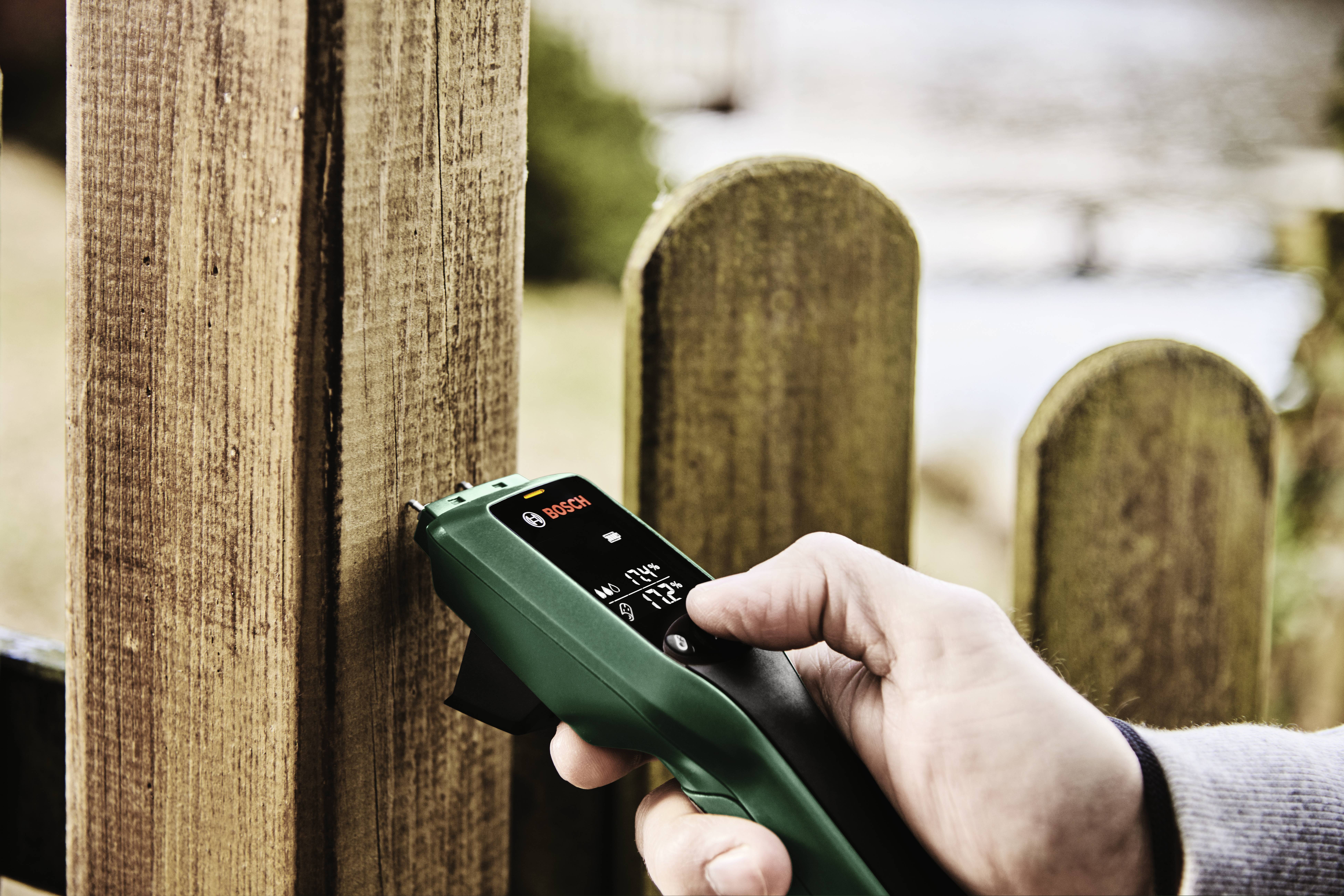 Une personne utilise un appareil de mesure sur une clôture en bois pour vérifier l'humidité du bois. Un jardin est visible de manière floue en arrière-plan.