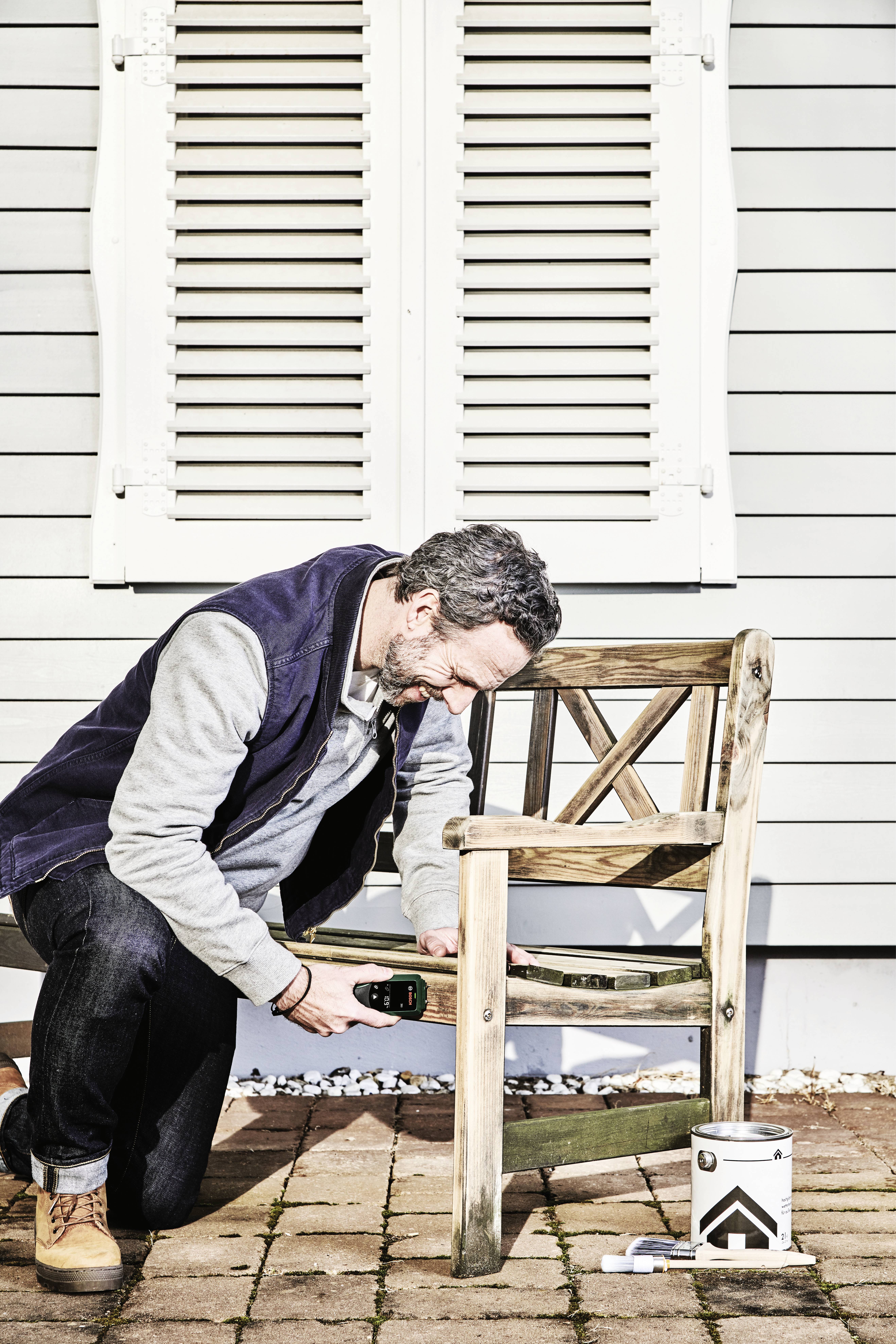 Un homme peint une table en bois à l'extérieur devant une maison blanche, tenant un pinceau, avec un pot de peinture posé à côté de lui.