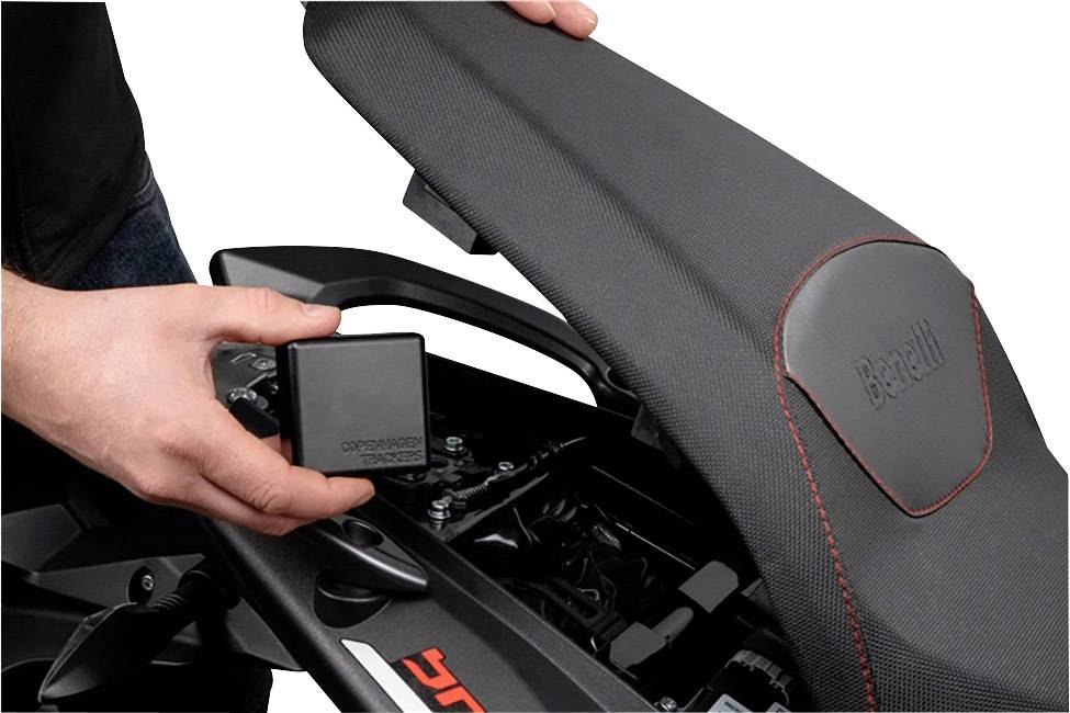 Une personne soulève la selle d'une moto pour montrer un petit appareil carré. La housse de selle est brodée du nom 'Benelli'.