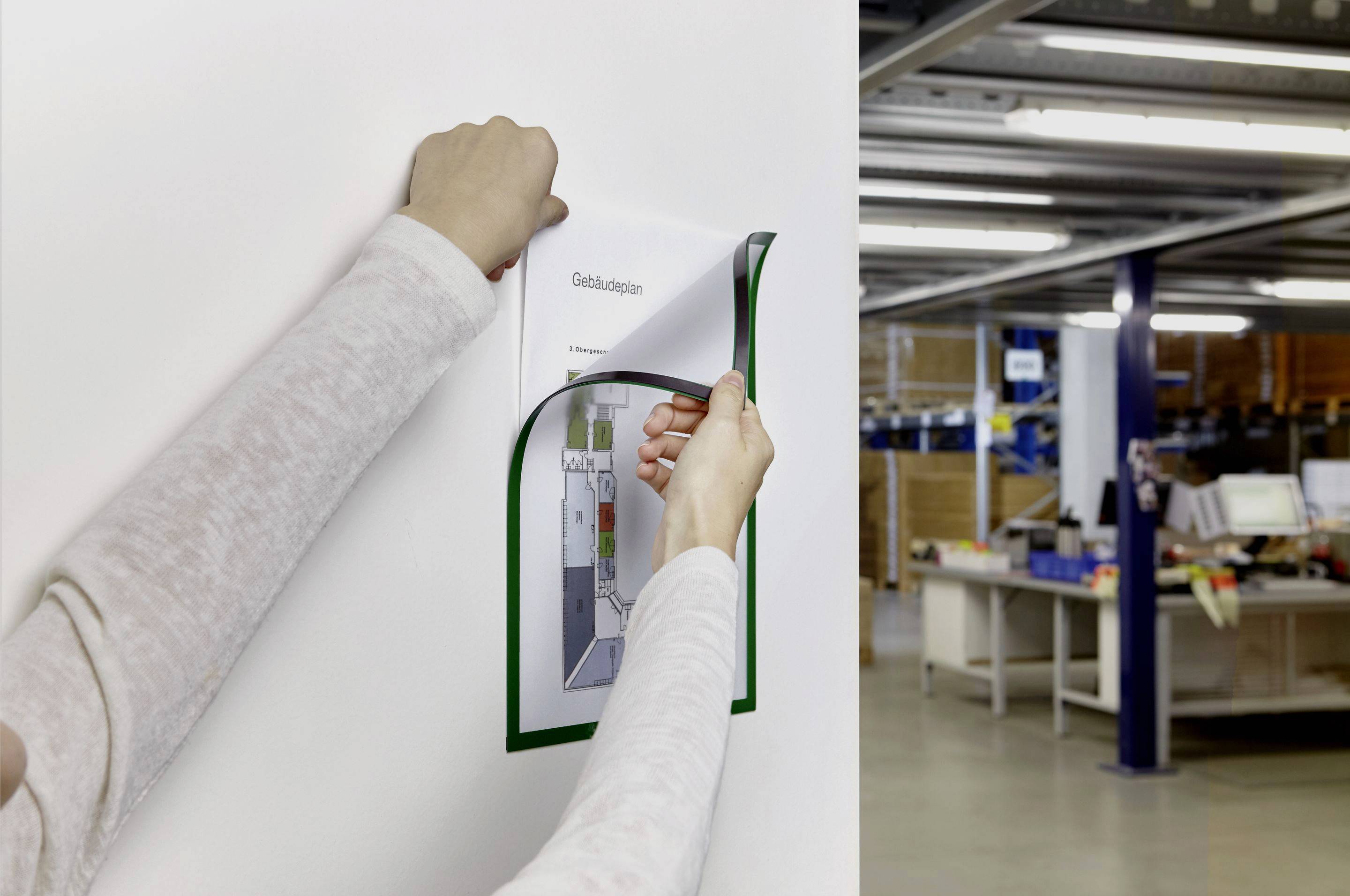 Une personne modifie un document sur un mur blanc dans un bureau. Des étagères et des postes de travail informatiques sont visibles en arrière-plan.